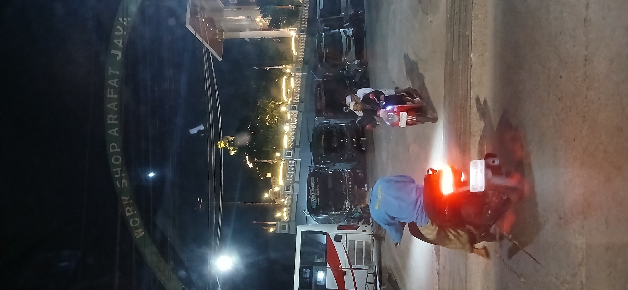 The image captures a nighttime scene at what appears to be a transportation hub or parking area. An arched sign overhead reads "WORK SHOP ARAFAT JAVA." In the background, a statue or monument with golden accents is visible, illuminated by string lights that also adorn a decorative railing and building facade.
In the foreground, a white bus with red and white stripes is parked on the left. Several dark-colored buses are parked behind it. Two men on a red scooter are riding towards the viewer. The driver is wearing a white cap and a dark shirt, while the passenger behind him is also wearing a white cap. Another person, seen from behind, is riding a dark motorcycle in the foreground, wearing a blue jacket and light-colored pants. The motorcycle's taillight is brightly illuminated, casting red light on the asphalt. The ground is paved with concrete, showing some cracks and texture. The overall atmosphere is dimly lit by artificial lights, suggesting it is a busy area even at night. The presence of buses, a workshop sign, and the general setting suggests this location could be a bus terminal, a depot, or a facility related to transportation in Samarinda, Indonesia.