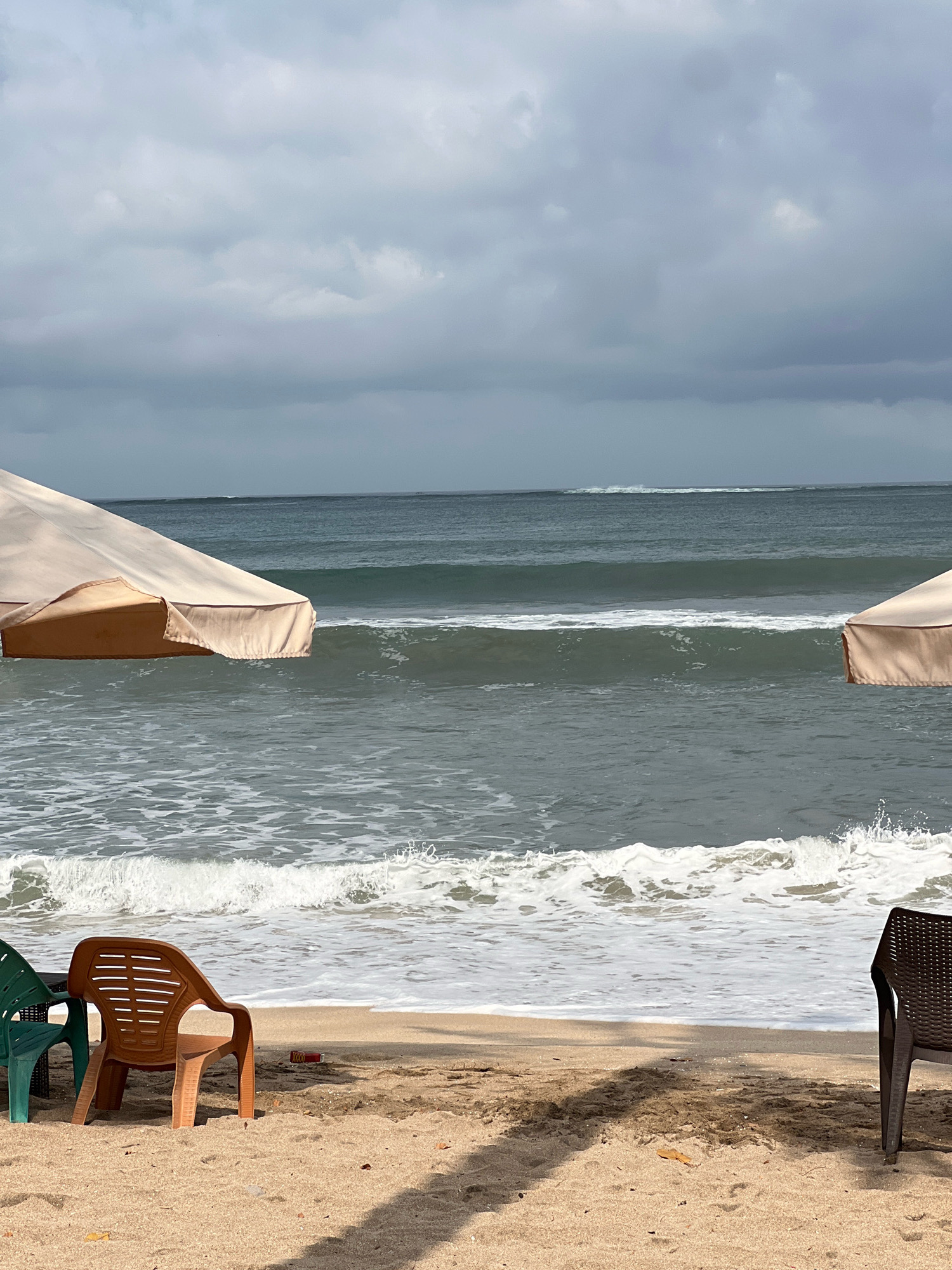 The image depicts a beach scene in Kuta, Indonesia, under a cloudy sky. In the foreground, on the sandy beach, are three plastic chairs: one green, one light brown, and one dark brown. A small red object, possibly a bottle or can, is visible on the sand between the chairs. Two large, beige beach umbrellas are positioned in the middle ground, one on the left and one on the right, casting shadows on the sand. The ocean is in the background, with moderately sized waves breaking and receding towards the shore. The water is a deep green-blue, and the sky is filled with overcast, gray clouds. The overall impression is one of a quiet, somewhat overcast day at the beach.