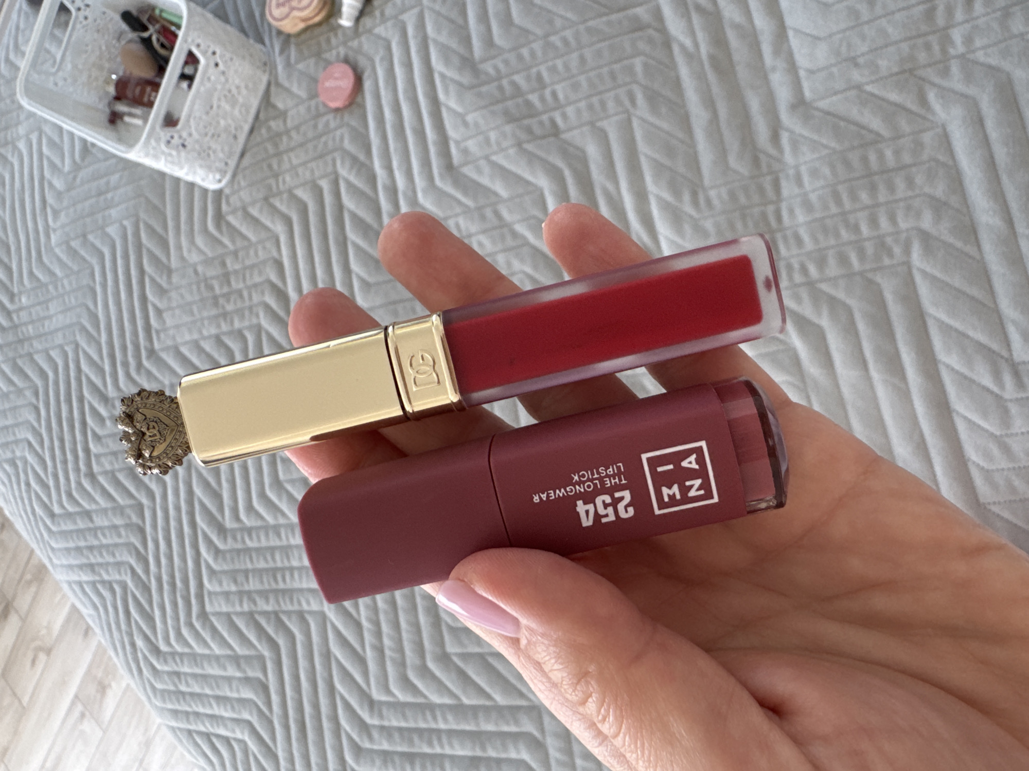 A close-up, overhead image captures a person's hand holding two distinct lip products against a textured background. The hand, featuring neatly manicured nails with a light pink polish, prominently displays the items.
The top product is a clear tube containing a vibrant red liquid lipstick. It has a glossy gold cap that features a "DG" logo, and one end of the cap is adorned with an intricate, crown-like emblem. Below it, the second product is a matte, rectangular lipstick tube in a plum or mauve tone. Visible text on this tube clearly reads "254," "THE LONGWEAR LIPSTICK," and a vertically arranged brand logo that appears to be "ZIA MZN."
In the blurred background, a light gray quilted blanket with a distinct geometric pattern covers a surface. To the upper left, a white perforated plastic basket holds various makeup items and brushes. A small, round, pink cosmetic compact is also visible on the blanket near the top right. A light-colored wooden floor is partially seen in the lower left corner of the frame.
The image suggests the activity of showcasing or selecting these cosmetic products. The indoor setting, likely a personal space such as a bedroom or dressing area, is illuminated by bright, diffused lighting, indicating it is daytime. The context provided indicates this scene is set in Sochi, Russia.