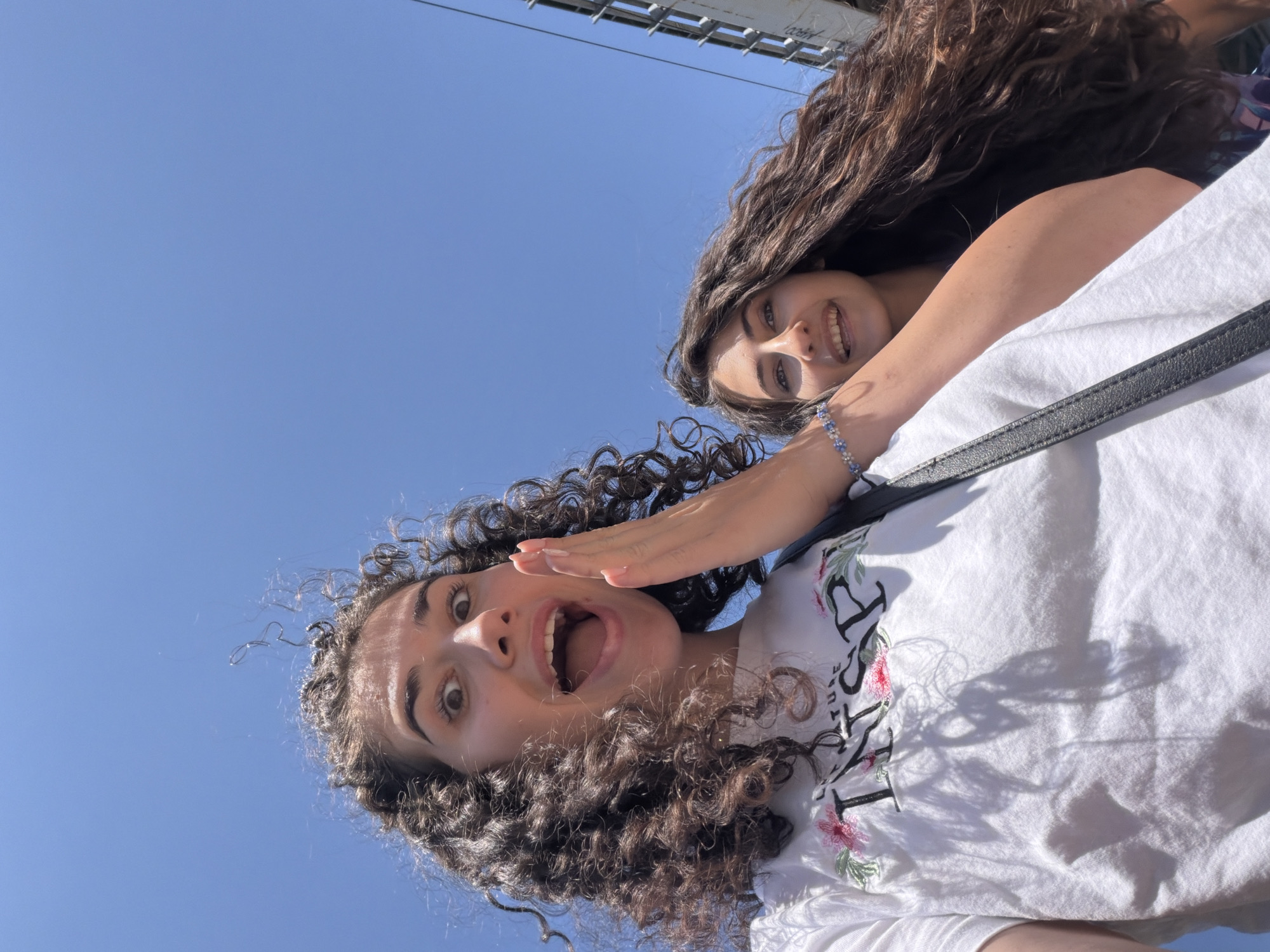 Two young women with long, dark, curly hair are posing for a selfie outdoors under a clear blue sky. The woman in the foreground, on the left, is looking directly at the camera with a surprised expression, her mouth open, and her hand raised to her cheek. She is wearing a white t-shirt with floral print and text that appears to say "INSPIRE." The woman in the background, on the right, is also looking towards the camera and smiling. She is wearing a bracelet on her left wrist. A black strap, likely from a bag, crosses the foreground woman's chest. A metallic structure, possibly a bridge or building railing, is visible on the right side of the image. The lighting suggests it is daytime. The image's metadata indicates it was taken in Sofia, Bulgaria, on May 27, 2024.