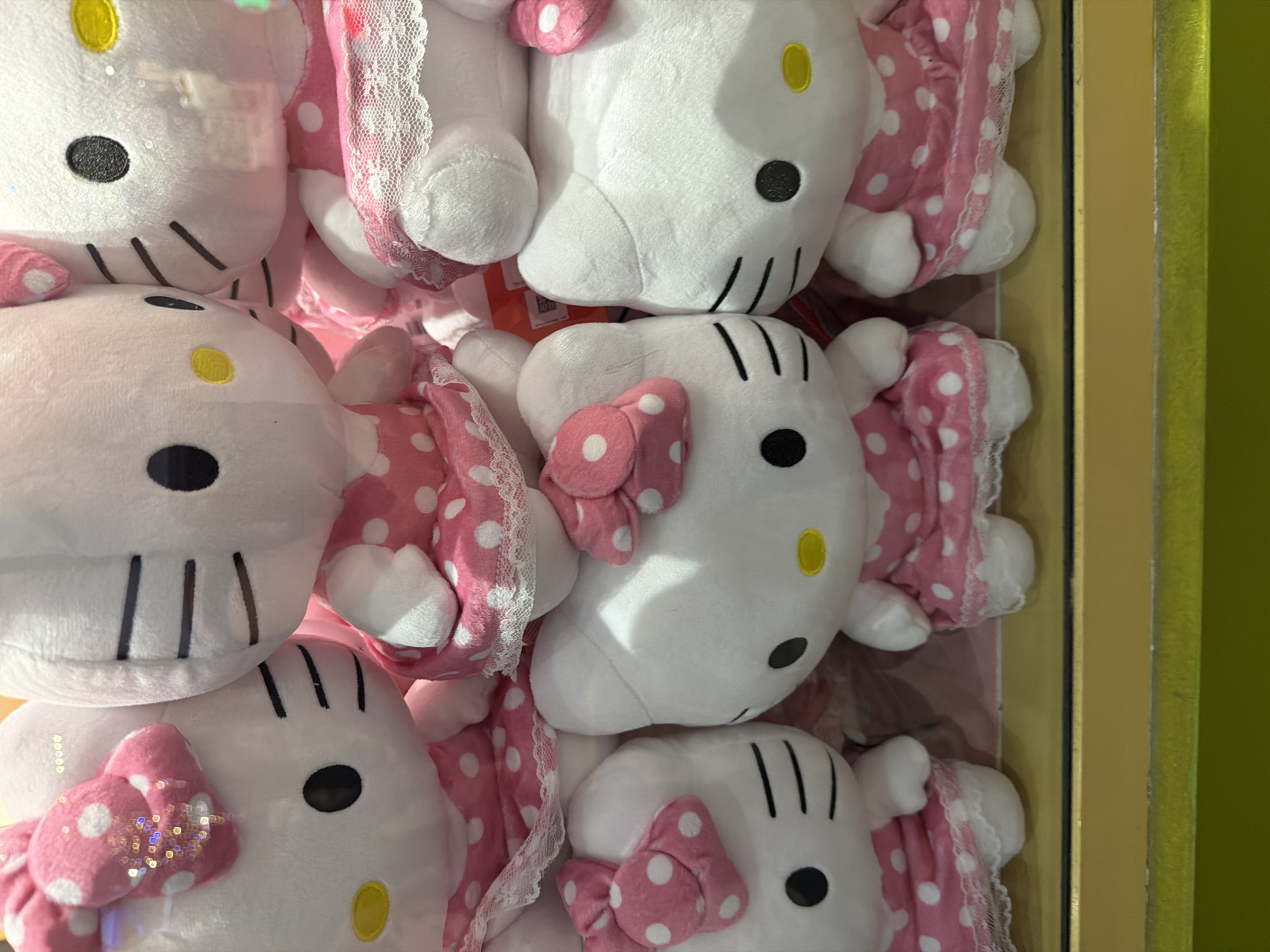A close-up, eye-level medium shot captures a stack of plush Hello Kitty toys, likely in a retail setting. The toys are predominantly white with pink polka-dot dresses. In the foreground, two Hello Kitty toys are visible side-by-side. The left toy faces forward with its head slightly tilted to the right, adorned with a large pink polka-dot bow on its head. Its dress has a lace trim. To its right, another Hello Kitty toy faces slightly left, also wearing a pink polka-dot dress with lace trim, and its bow is partially obscured. Behind these, more Hello Kitty toys are stacked, with varying degrees of visibility. The lighting is bright, highlighting the textures of the plush toys and the details of their clothing. The background is blurred, suggesting a shallow depth of field. The scene is framed by a golden-yellow border at the bottom, with a hint of a green surface below that.