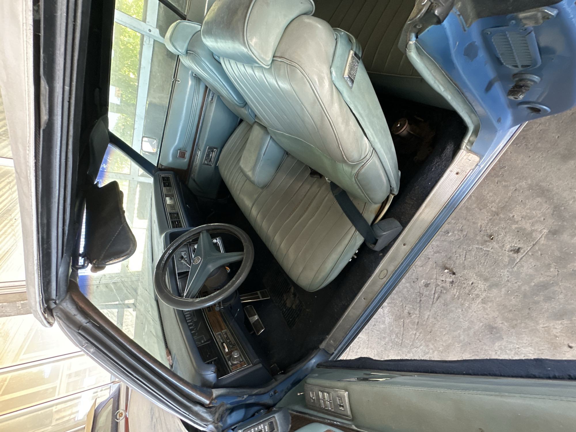 An interior view of a classic blue convertible car. The vehicle is parked indoors, possibly in a garage or under a covered structure. The light-colored interior features light blue vinyl seats with a fold-down armrest between the front seats. A dark steering wheel is visible in the foreground, along with the dashboard and radio. The convertible top is up. The car's door is open, revealing the interior and the concrete floor outside.