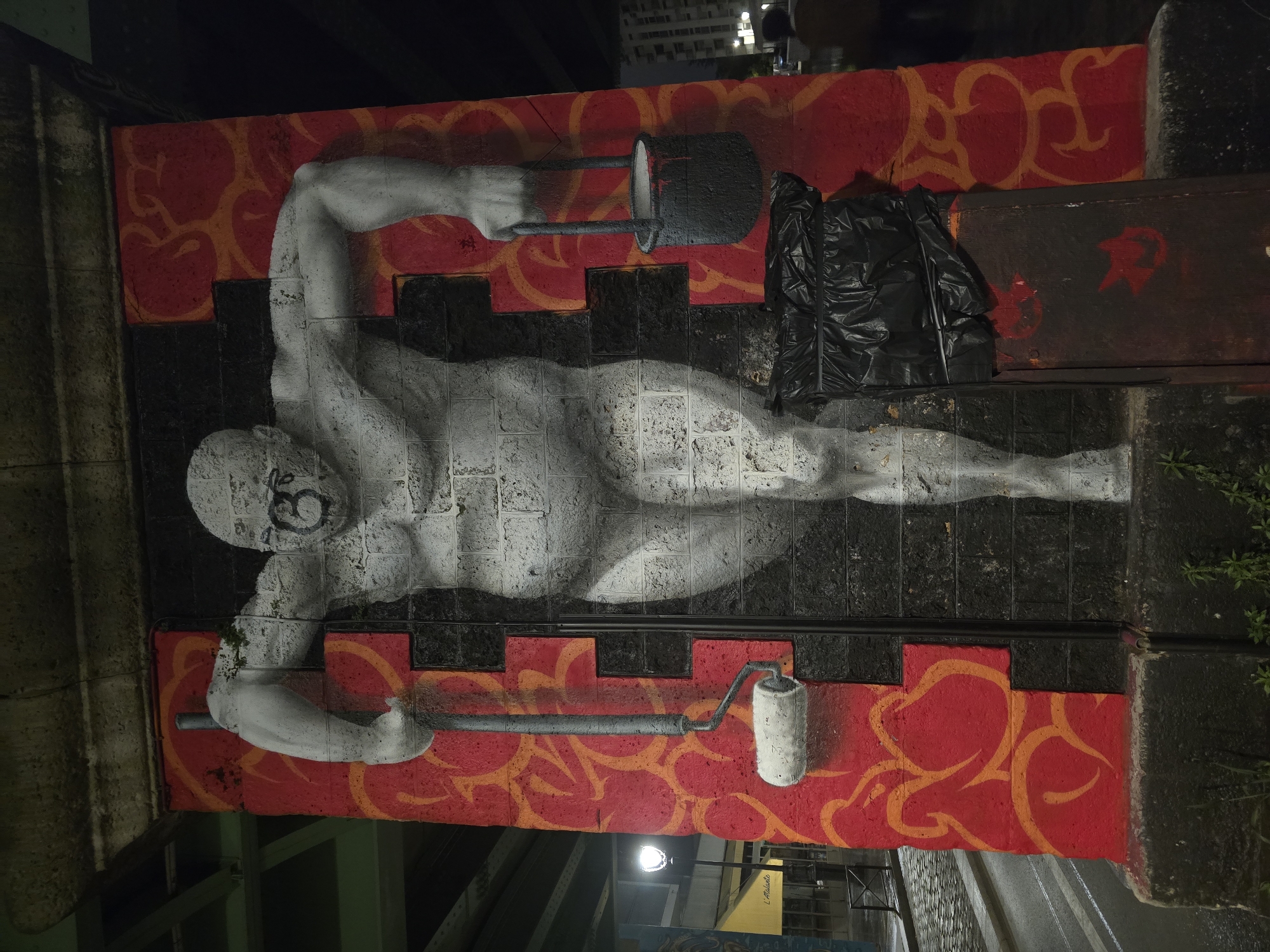Captured at night, the image showcases a mural in what appears to be Paris, France. The mural, situated on a horizontal concrete wall or barrier, features a stylized rendition of a muscular figure in grayscale, depicted as if lying on its side. The figure is holding painting tools: a paint roller in one hand and a bucket of paint in the other.
The mural is painted against a backdrop of red with orange swirling patterns above and below the figure, with black rectangular blocks running through the middle. Next to the figure, a black trash bag and a rusty metal sheet adorned with red graffiti are positioned on the wall. The overall environment surrounding the mural appears urban and slightly gritty, with glimpses of surrounding infrastructure, including possible streetlights and foliage.