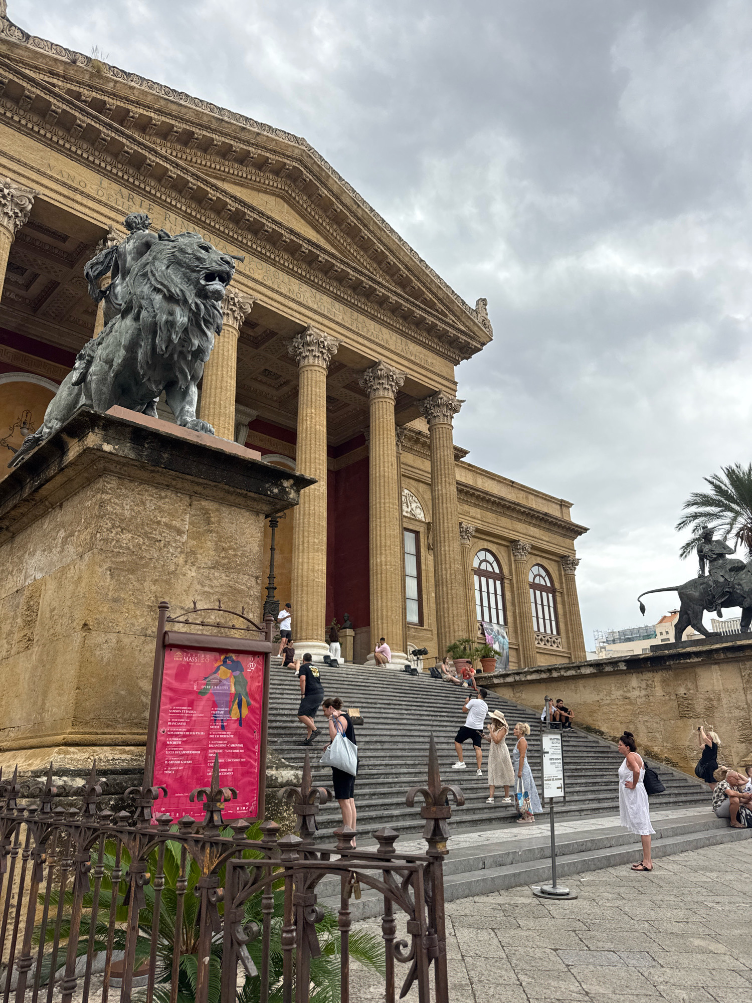 The image depicts the Teatro Massimo in Palermo, Italy, a grand opera house renowned for its Neoclassical architecture. The exterior features a prominent facade with towering Corinthian columns, ornate friezes, and a triangular pediment. In the foreground, a formidable bronze lion statue stands guard on a stone pedestal, its mane elaborately detailed. A wide, imposing staircase leads up to the opera house entrance, where several people are visible.
The staircase is a central element of the scene, with individuals in various poses: some ascend, others descend, and a few are paused, possibly taking in the sights or waiting for companions. Among the people are tourists and locals, dressed in casual summer attire. One woman in a white dress stands near the bottom of the stairs, holding a tote bag. Further up, a couple in light-colored clothing walks together, and a man in a black t-shirt and shorts is captured mid-stride. On the left side of the stairs, a group of individuals is seated, enjoying the day.
A large red poster, likely advertising an event or opera, is affixed to a metal stand to the left of the staircase, its text partially obscured but clearly visible enough to identify it as a cultural announcement. The overall atmosphere is one of a lively, public space on a cloudy day, suggesting a popular tourist destination. The architecture of the Teatro Massimo dominates the background, with its imposing scale and historical significance evident. To the right, another bronze sculpture of a lion is visible, further emphasizing the iconic imagery associated with the opera house. The sky is overcast, casting a diffused light over the scene.