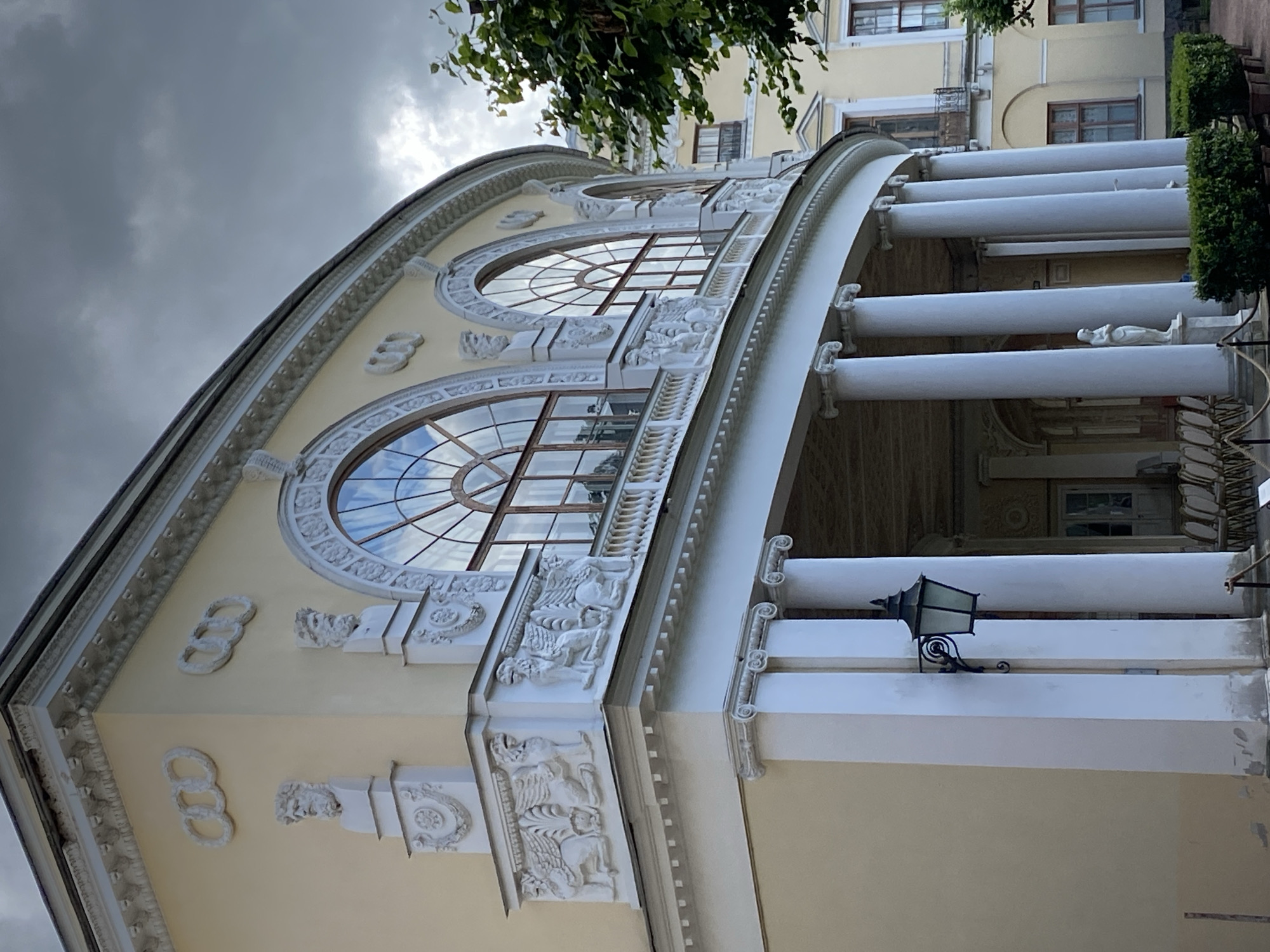 This is a low-angle exterior, eye-level shot of the rounded facade of a yellow building, likely the Pavlovsk Palace or an adjacent structure in Pavlovsk, Russia. The architecture features classical elements, including a colonnade of white, fluted columns supporting a curved portico. Above the portico, large arched windows with intricate mullions reflect a cloudy, overcast sky.
Decorative elements are prominent:
* On the upper facade, there are circular relief sculptures, each composed of three interlocking rings.
* Below these, busts of bearded men are mounted on pedestals, flanking the windows.
* Further down, on a projecting ledge, white relief sculptures of winged lions or griffins are depicted in profile, seated with their forelegs extended.
* The balustrade along the upper level is also ornate.
* A black, wrought-iron lantern with a glass enclosure hangs from the wall near the left side of the image, attached to one of the columns.
In the foreground, neatly trimmed green bushes in planters are visible along the edge of the portico. A white statue of a robed female figure stands on a pedestal within the portico area. Chairs are arranged in rows behind the bushes, suggesting an outdoor seating area. A tree with green foliage is present on the right side of the frame, partially obscuring the building. The overall impression is one of grand, historical architecture under a dramatic, cloudy sky.