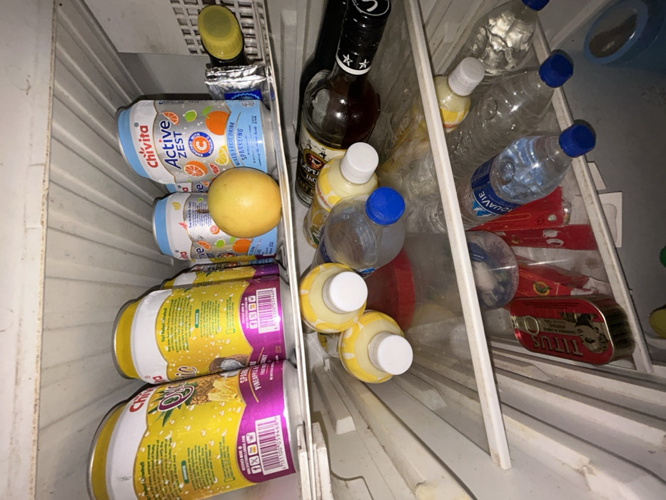 The image displays the interior of a refrigerator, located in Egbe, Nigeria, containing multiple food and beverage items distributed across three visible shelves. The top shelf holds several 330ml cans of Chivita 100% Sparkling Pineapple Fruit Drink and Chivita Active Zest Mixed Fruit Drink Sparkling, along with a single yellow, spherical fruit resembling a grapefruit. A dark-colored bottle with a yellow cap is partially visible on the far right of this shelf. The middle shelf contains several small plastic bottles filled with a yellow-white liquid, numerous clear plastic water bottles (including one labeled "AQUAVIE"), and a dark glass bottle with a black cap and white star emblem positioned horizontally. A red plastic container and various plastic wrappers are also present. The bottom shelf, partially visible, features a red rectangular tin of Titus Sardines and additional plastic bottles. The refrigerator's interior walls and shelves appear worn, with some visible grime.