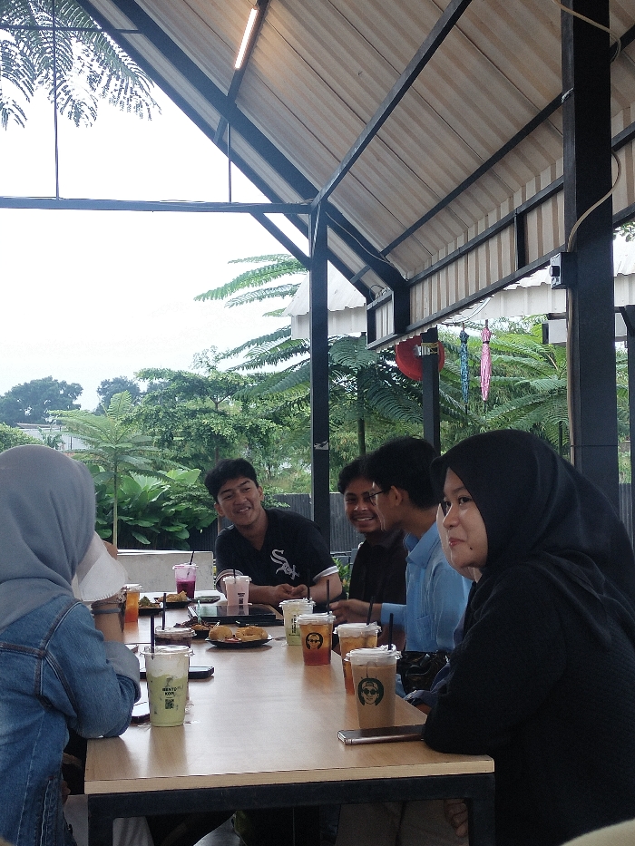 The image displays a group of young people socializing at an outdoor cafe or restaurant in Dramaga, Indonesia. Five individuals are seated around a light wooden table, enjoying drinks and food.
On the left, a person wearing a light blue hijab and a denim jacket is seen from behind. Next to them, a young man in a black t-shirt, featuring a "WHITE SOX" logo, is smiling and looking towards the left. Beside him, another young man with glasses is also smiling, looking towards the right. A fourth person, wearing a light blue collared shirt, is partially visible, facing right. On the far right, a woman in a black hijab and black top is seated, facing right, with a slight smile.
The table is filled with several clear plastic cups containing various beverages, such as what appears to be matcha (with "BENTO CHA" visible on one cup), coffee, and other colorful drinks, all with straws. Many cups feature cartoon-like faces printed on them. Plates with food, including fried items, are also on the table, along with mobile phones.
The setting is an open-air area with a sloped metal roof structure composed of black beams and corrugated metal sheets, under which a light fixture is visible. The background reveals lush green foliage and trees under an overcast or cloudy sky, indicating daytime or late afternoon. Two colorful umbrellas, one pink and one blue, hang decoratively in the background. The smiling expressions on the faces of the people suggest a cheerful and relaxed gathering among friends.