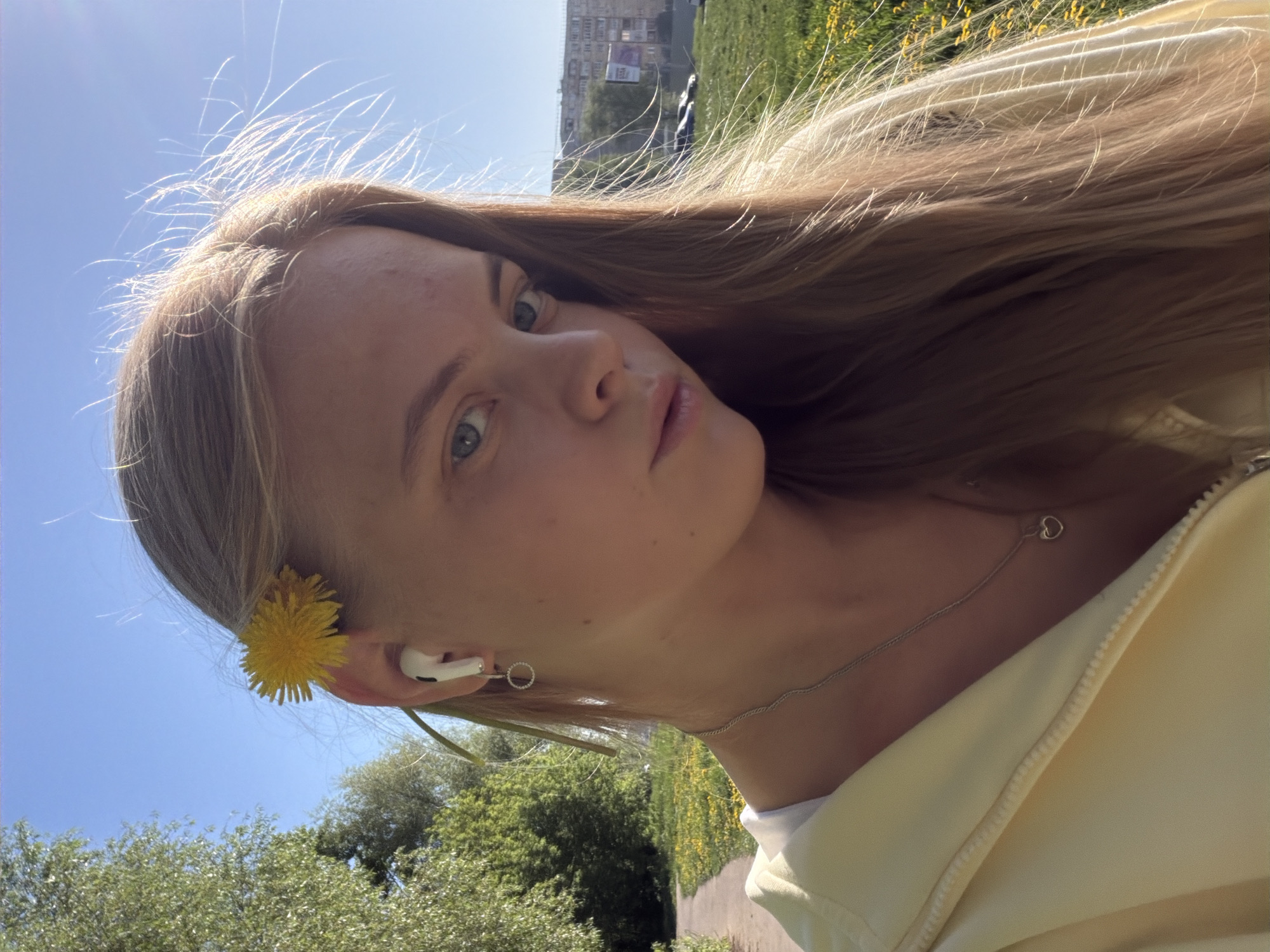 A young, fair-skinned woman with long blonde hair is the subject of a selfie. She has blue eyes and a neutral expression. A yellow dandelion is placed behind her left ear, next to an earpod. She is wearing a silver necklace with a small heart pendant and a light yellow jacket. The background includes a blue sky, green trees, and a grassy area with dandelions. In the distance, there is a building that appears to be located in an urban area. The location is Izhevsk, Russia.