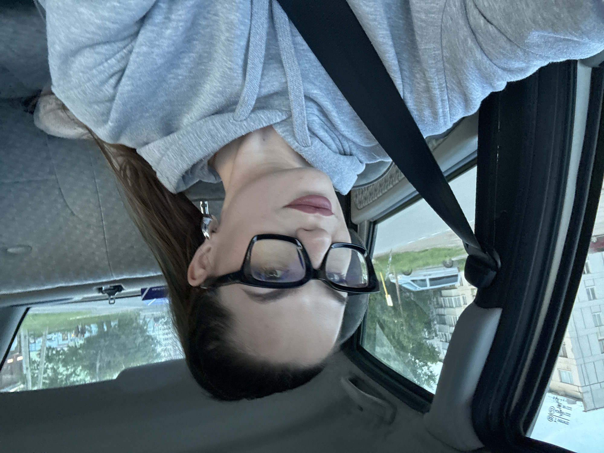 A medium shot shows a woman with dark hair pulled back, wearing large black-rimmed glasses and a grey hooded sweatshirt, seated in the passenger seat of a car. Her seatbelt is fastened across her body. The interior of the car is visible, including the grey fabric of the seats and the car's ceiling. Through the open car window, an urban landscape is depicted, featuring multi-story residential buildings with light-colored facades and balconies, lush green trees, and a street with a silver van parked on the side. The scene appears to be in Chaikovsky, Russia, given the context of the provided information. The lighting suggests it is daytime, possibly late afternoon or early evening.