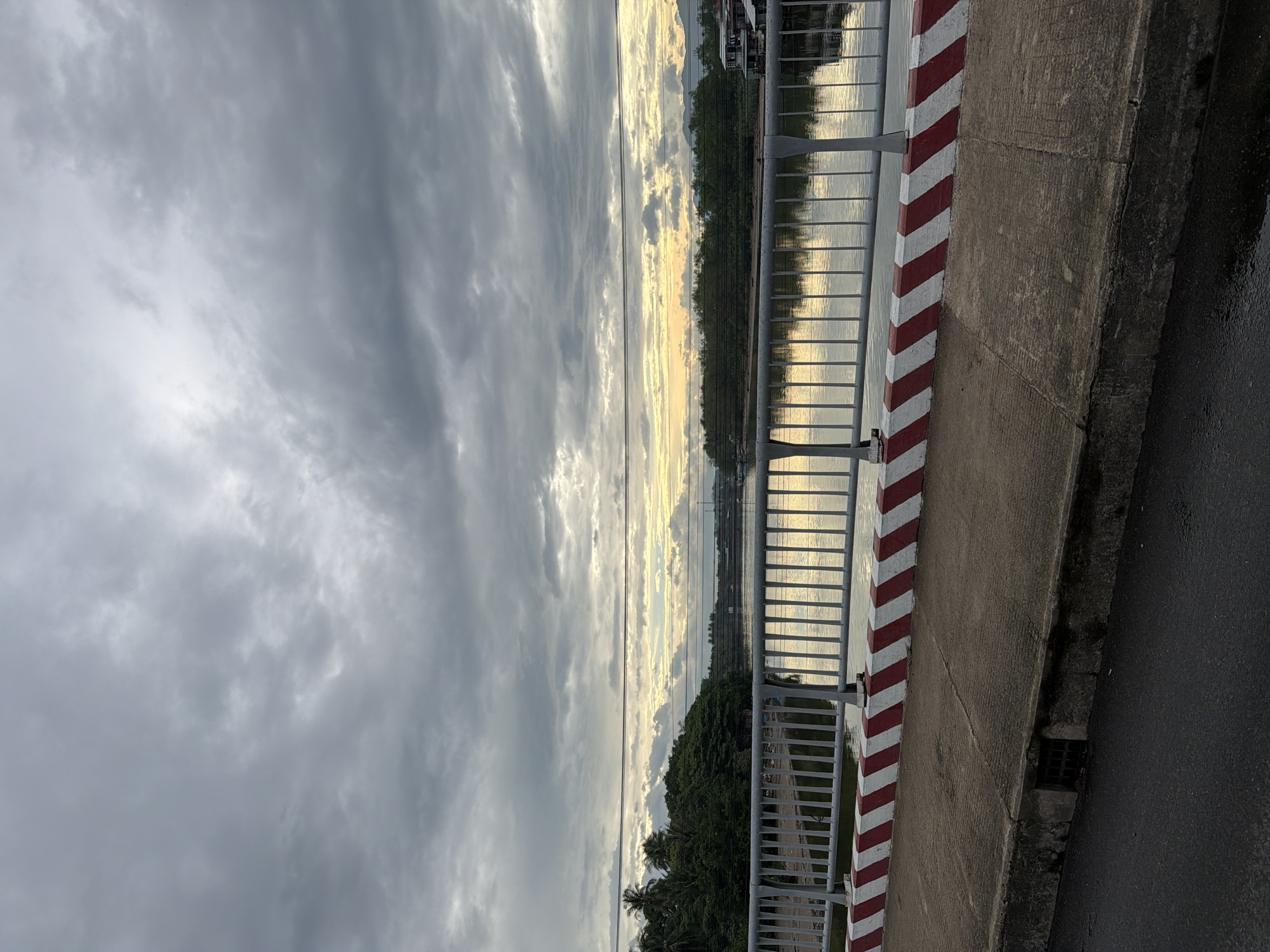 A vertical, medium shot captures a bridge railing overlooking a calm river and a tree-lined shore under a cloudy sky. The bridge railing is made of silver-colored metal bars with a white and red diagonal stripe pattern painted on the concrete barrier below. The river reflects the muted light from the overcast sky and a sliver of yellow sunset peeking through the clouds on the horizon. Dense green trees with palm fronds line the opposite bank, with hints of structures and distant mountains visible beyond. Power lines stretch horizontally across the upper portion of the frame, obscuring a clear view of the sky. The foreground shows the concrete walkway of the bridge and a portion of the asphalt road, with a drain grate visible on the side. The overall scene depicts a serene, possibly pre-dusk, moment in a coastal city. Based on the visual cues of lush tropical vegetation, waterfront structures, and the overall ambiance, this scene is consistent with locations in Nha Trang, Vietnam.