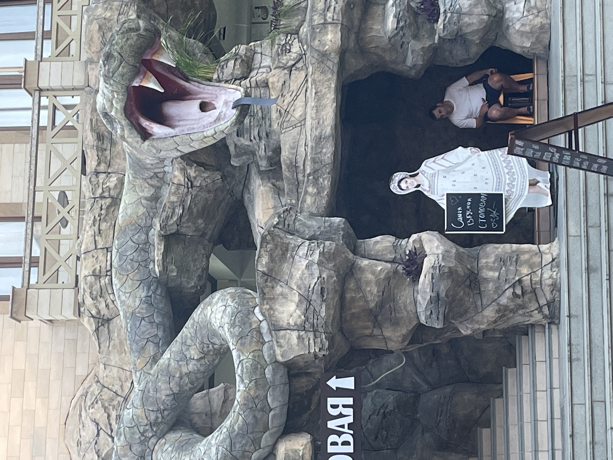 An outdoor scene shows a large, sculpted snake emerging from a rock formation that resembles a cave. The snake's mouth is wide open, revealing a red interior and sharp white fangs. A dark blue object hangs from its mouth, resembling a tongue or a stream of water. The rock formation has a rough, textured surface, with cracks and crevices.
In front of the rock formation, on a raised platform with stairs leading up to it, are two figures. To the left is a life-sized cardboard cutout of a woman wearing a traditional white embroidered dress and a headpiece. She holds a blackboard with text written in what appears to be Cyrillic. To the right of the cutout, a man is seated on a small orange chair. He is wearing a white t-shirt and dark shorts. He appears to be looking to his left.
A dark brown A-frame sign with a menu is positioned in the foreground, angled towards the right. Above the rock formation, a building with windows and a decorative railing is visible. The overall impression is of an amusement or tourist attraction.