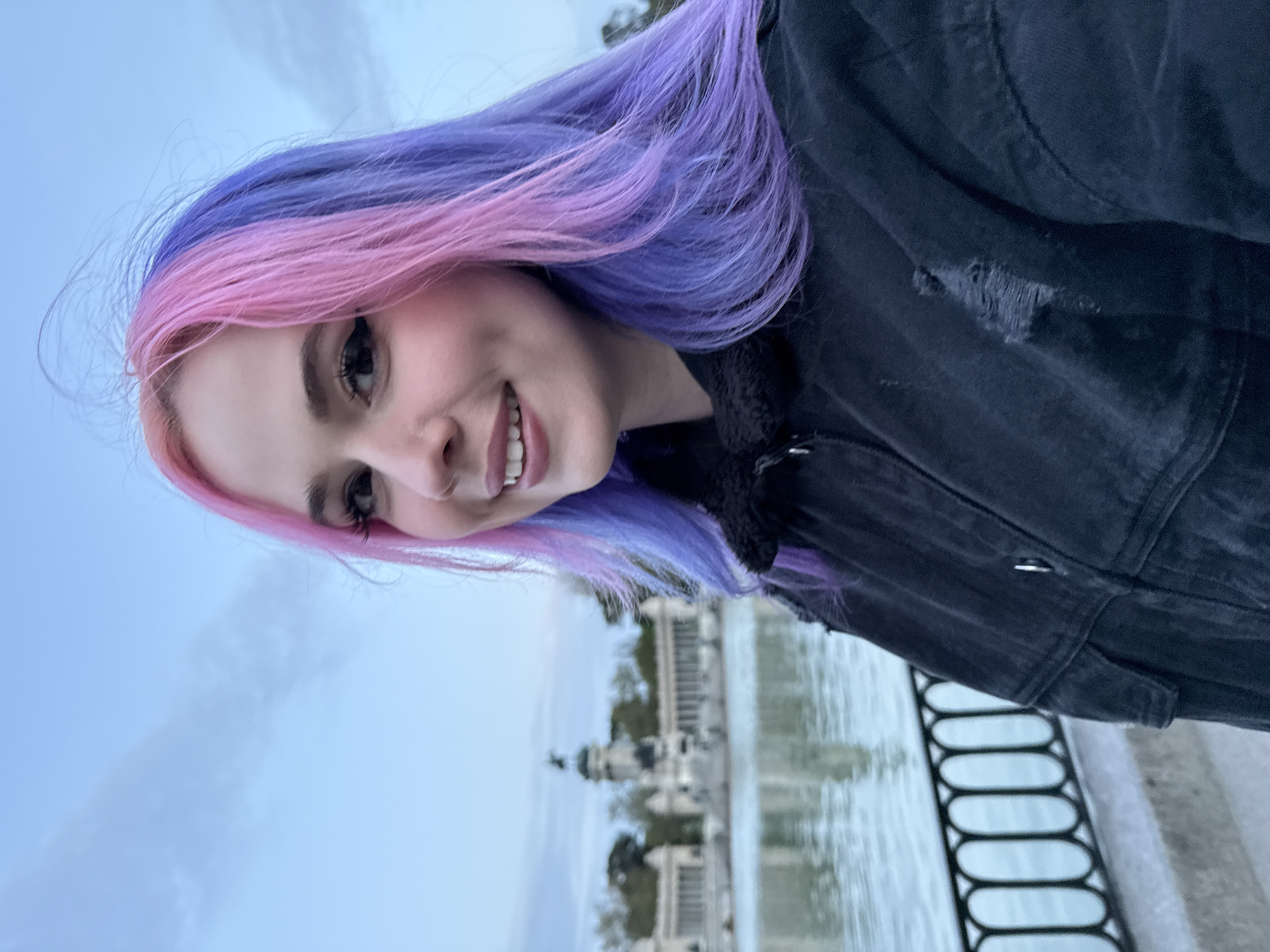A selfie shows a smiling young woman with pink and purple hair standing near the Estanque del Retiro in Madrid, Spain. The Estanque's monument is partially visible in the background, reflected in the water. A black jacket with a ripped section is visible on the right side of the frame. The sky is overcast. A portion of a black metal fence runs along the bottom right.
