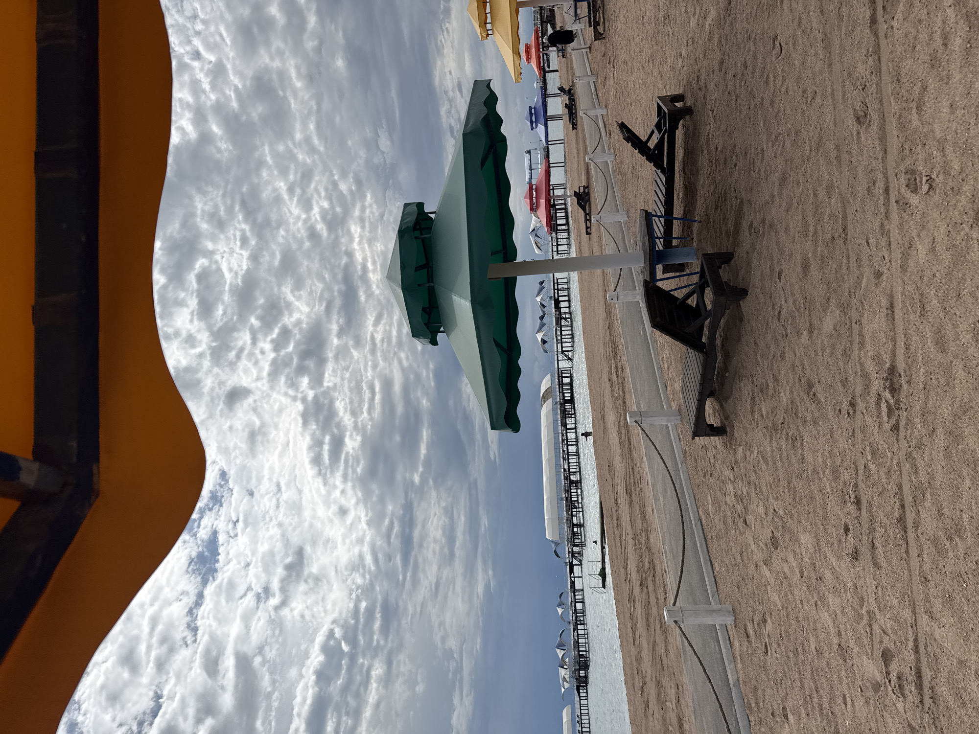 This is a photograph of a beach scene in Sary-Oy, Kyrgyzstan, under a cloudy sky. The foreground is dominated by tan sand with some footprints. Two dark wooden lounge chairs are placed on the sand, one angled towards the right and the other towards the left. In front of the left lounge chair is a small blue table with a blue leg.
Further back, a path runs horizontally across the sand, marked by white posts and a dark rope. Beyond the path, several beach umbrellas are visible in a variety of colors: a large green umbrella is prominently featured on the right, with yellow, red, and blue umbrellas further down the beach. Some of these umbrellas cover more lounge chairs.
The middle ground features the shore of a body of water, appearing calm and with ripples. In the distance, a long pier stretches out over the water, supported by numerous pilings. Several white tent-like structures with arched roofs are situated along this pier. A few people can be seen in the water near the pier, and one person is standing on the pier.
The background is filled with a dramatic, cloudy sky with patches of lighter and darker grey clouds, suggesting overcast or potentially stormy weather. An orange-colored structure, possibly part of a canopy or roof, partially frames the top of the image.