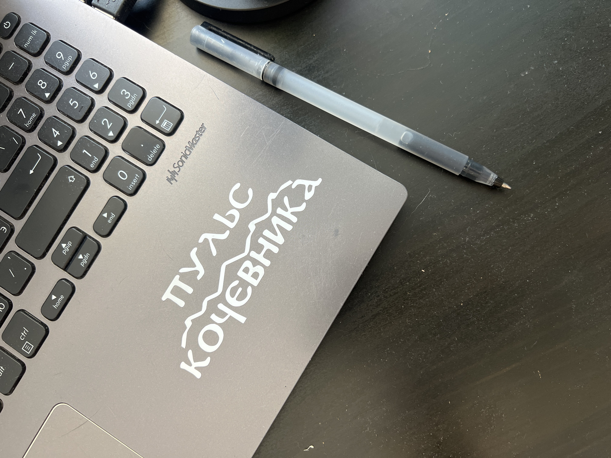 A close-up, overhead shot shows a portion of a silver laptop keyboard and a black wooden desk. A white sticker with Cyrillic lettering, "ПУЛЬС КОЧЕВНИКА," is affixed to the laptop. To the right of the laptop, a clear plastic pen with a black clip rests on the desk. The keyboard has various keys labeled, including "ctrl," "home," "pgup," "pgdn," "end," "insert," and "delete." The desk appears dusty. The scene is set in Novosibirsk, Russia.