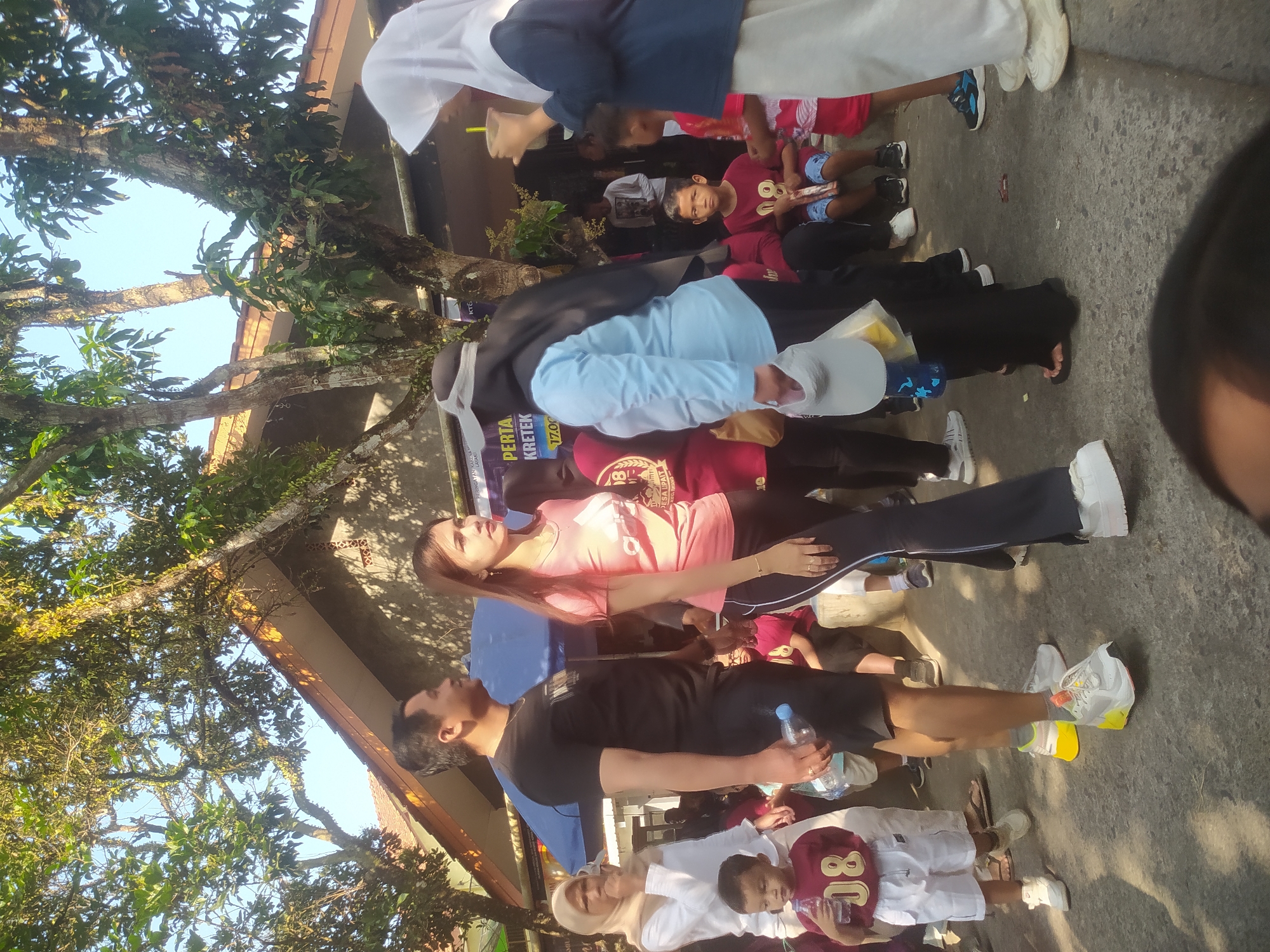 In Lopait, Indonesia, an outdoor gathering is underway on a paved surface, likely a street or community area, under a bright, sunny sky with dappled sunlight filtering through the leaves of overhead trees. Several individuals are present, engaged in various activities. A man in a black t-shirt and shorts holds a clear plastic bottle of water. Beside him, a woman in a pink t-shirt and dark pants stands attentively. In the background, a person in a light blue shirt and dark headscarf is seen interacting with someone holding a drink. Further back, a young boy in a maroon shirt with the number "08" is holding a bottle of water. Other people, including children and adults, are visible in the middle and background, some seated and others standing. A blue umbrella provides shade for some of the attendees. Banners with text are mounted on a building in the background, suggesting an organized event. The scene appears to be a community event or festival in Lopait.