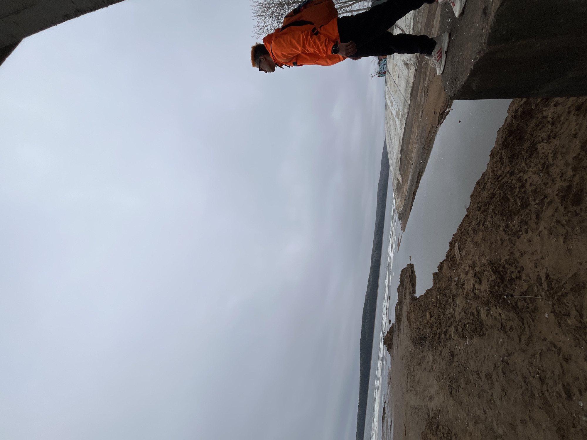 The image shows a man in an orange jacket standing on a concrete structure, possibly a pier or embankment, at a location near a body of water. The location is likely in the city of Chaikovsky, Russia. The man is wearing dark pants and light-colored shoes with stripes. The environment includes a beach, a body of water with some ice floes, and a cloudy sky. Some trees are visible in the background. The image is rotated 90 degrees counter-clockwise.