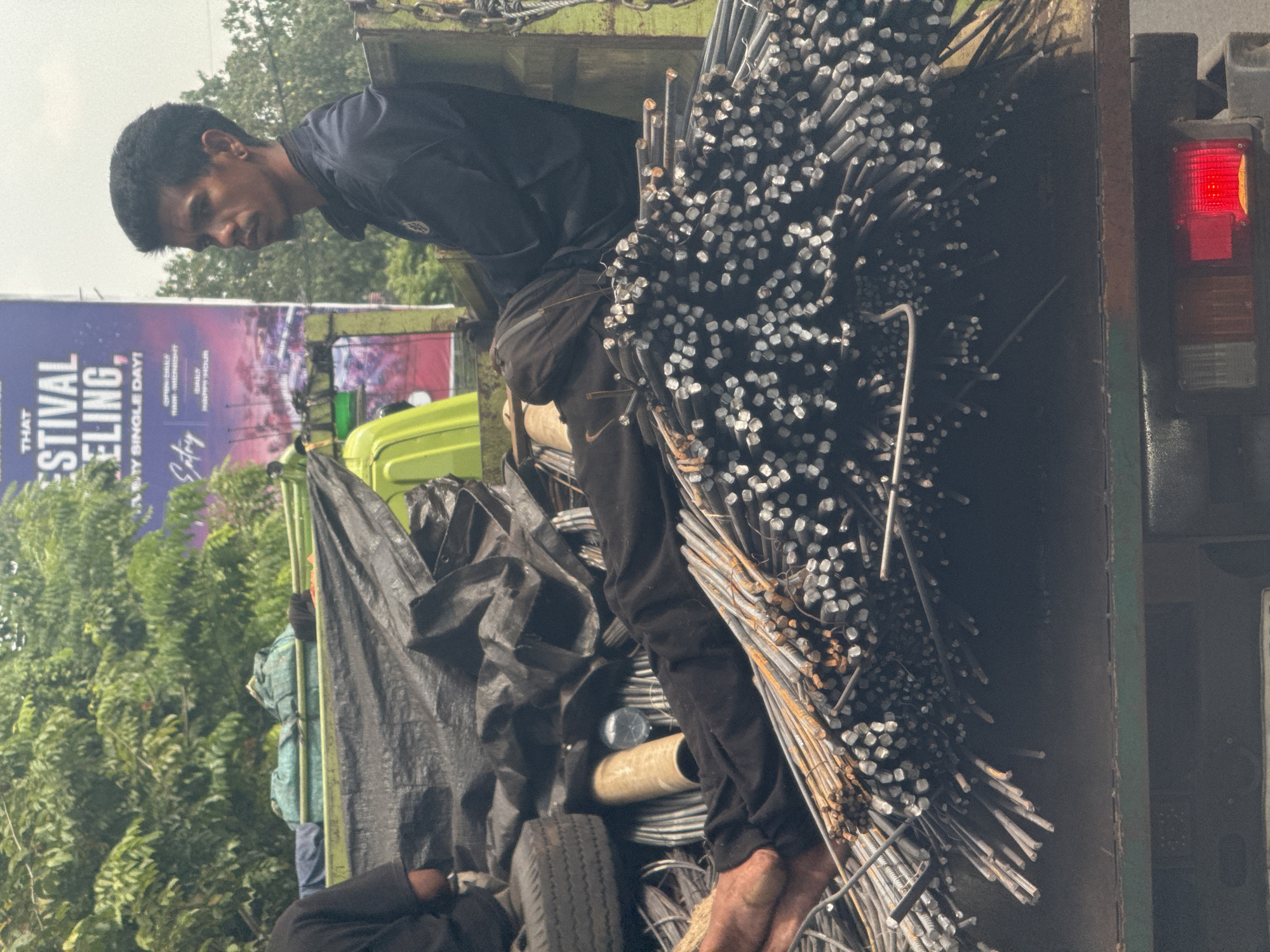 In North Kuta, Indonesia, an adult male stands barefoot inside the bed of a truck, amidst a large load of metal rods. He is clad in dark clothing. The truck bed also contains a tire, covered objects, and a tarp. A billboard featuring text "Festival Feling" is partially visible in the background, which includes lush green trees. A truck taillight is prominent on the right.