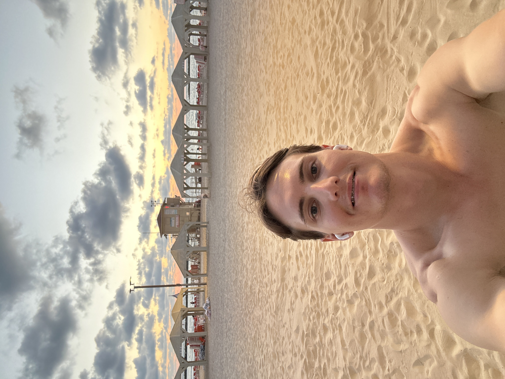 A medium shot depicts a shirtless man with short brown hair and earbuds, facing the camera in a selfie. The background reveals a sandy beach adjacent to a line of open-air shelters. In the distance, structures are visible on the beach, suggesting the beach is in Tel-Aviv, Israel. The sky displays a mixture of clouds and sunlight.