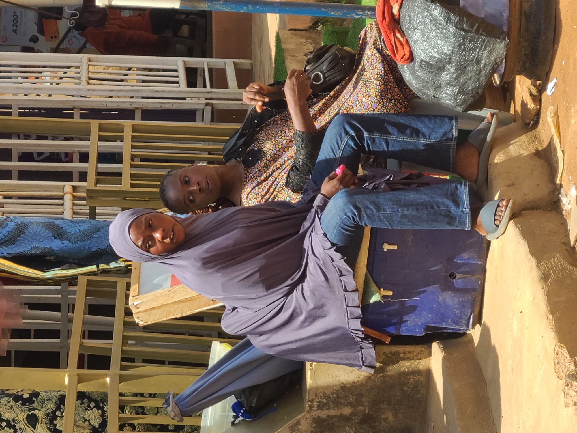 Two young women are seated outdoors in an urban environment, likely in Jos, Nigeria, as suggested by the architectural elements and the presence of a nearby market stall indicated by a sign with "A 200+" visible. The woman on the left is wearing a purple hijab that drapes over her shoulders and a blue denim outfit. She is seated on a blue plastic container. The woman on the right is wearing a vibrantly patterned dress and has a black handbag slung over her shoulder. She is looking to her left, with her hands engaged with her bag. They are sitting on a raised concrete structure. In the background, there is a metal fence and what appears to be merchandise displayed in a stall. The ground is dirt and sand, with some scattered debris.