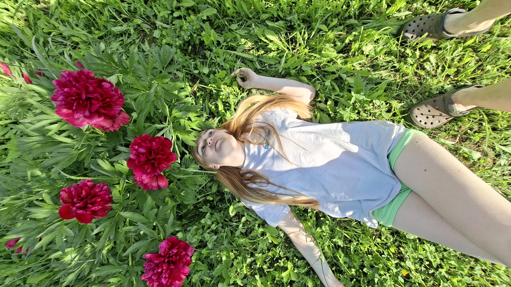 A young girl with long, blonde hair lies on her back in a lush green meadow dotted with bright pink peonies. She is wearing a light-colored t-shirt and green shorts. Her eyes are open, and she appears relaxed and content, gazing upwards. The sun is shining brightly, casting dappled shadows on the grass, suggesting it is a warm, sunny day, likely late morning or early afternoon. The setting is a natural outdoor environment, possibly a garden or a field, indicated by the abundance of grass and flowers. There are also two pairs of dark, casual clogs visible in the top right and bottom right of the frame, suggesting another person is nearby, perhaps taking the photo or also relaxing in the meadow. The overall mood of the image is peaceful and idyllic.