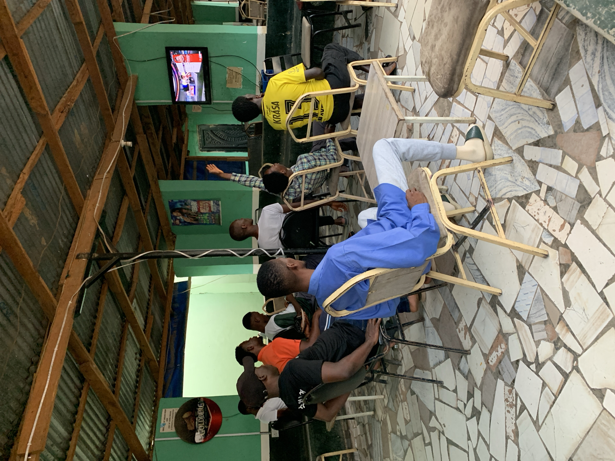 In a building in New Stadium, Nigeria, a group of people are seated on chairs facing a television screen mounted on a green wall. The building has a wooden support structure and a mosaic tile floor. The individuals are casually dressed, some in t-shirts and others in shirts. They appear to be watching something on the television. The environment suggests a public or semi-public gathering place.