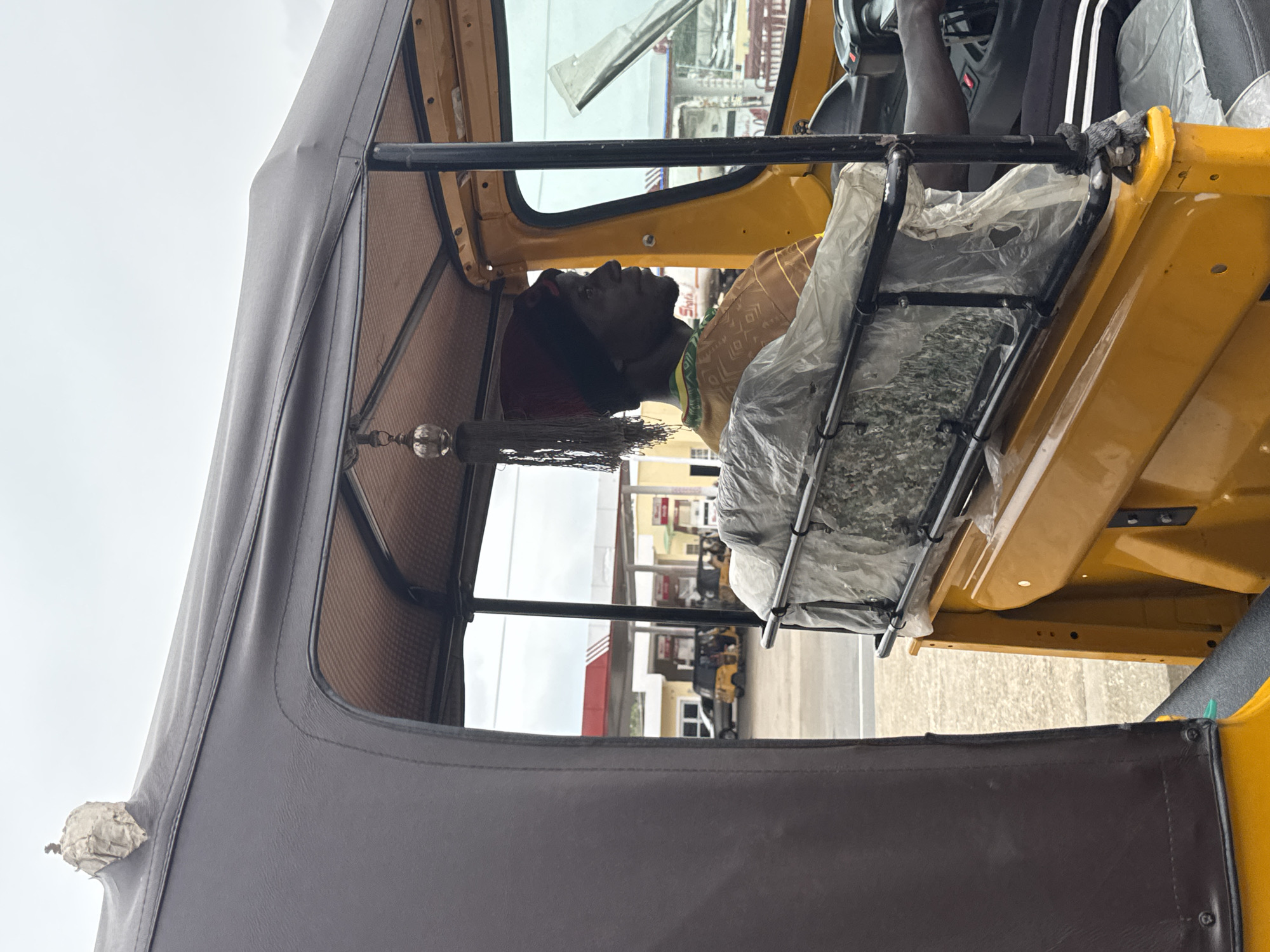 In the city of Jalingo, Nigeria, a man drives a yellow three-wheeled auto-rickshaw under a cloudy sky. The vehicle has a dark fabric roof and a decorative tassel hangs from the ceiling. The driver wears a red cap and a patterned brown and yellow shirt with green trim. His right hand is on the steering wheel. In the background, a gas station with red and white signage and several other vehicles are visible. The auto-rickshaw's interior has a black metal frame supporting a plastic-covered seatback.