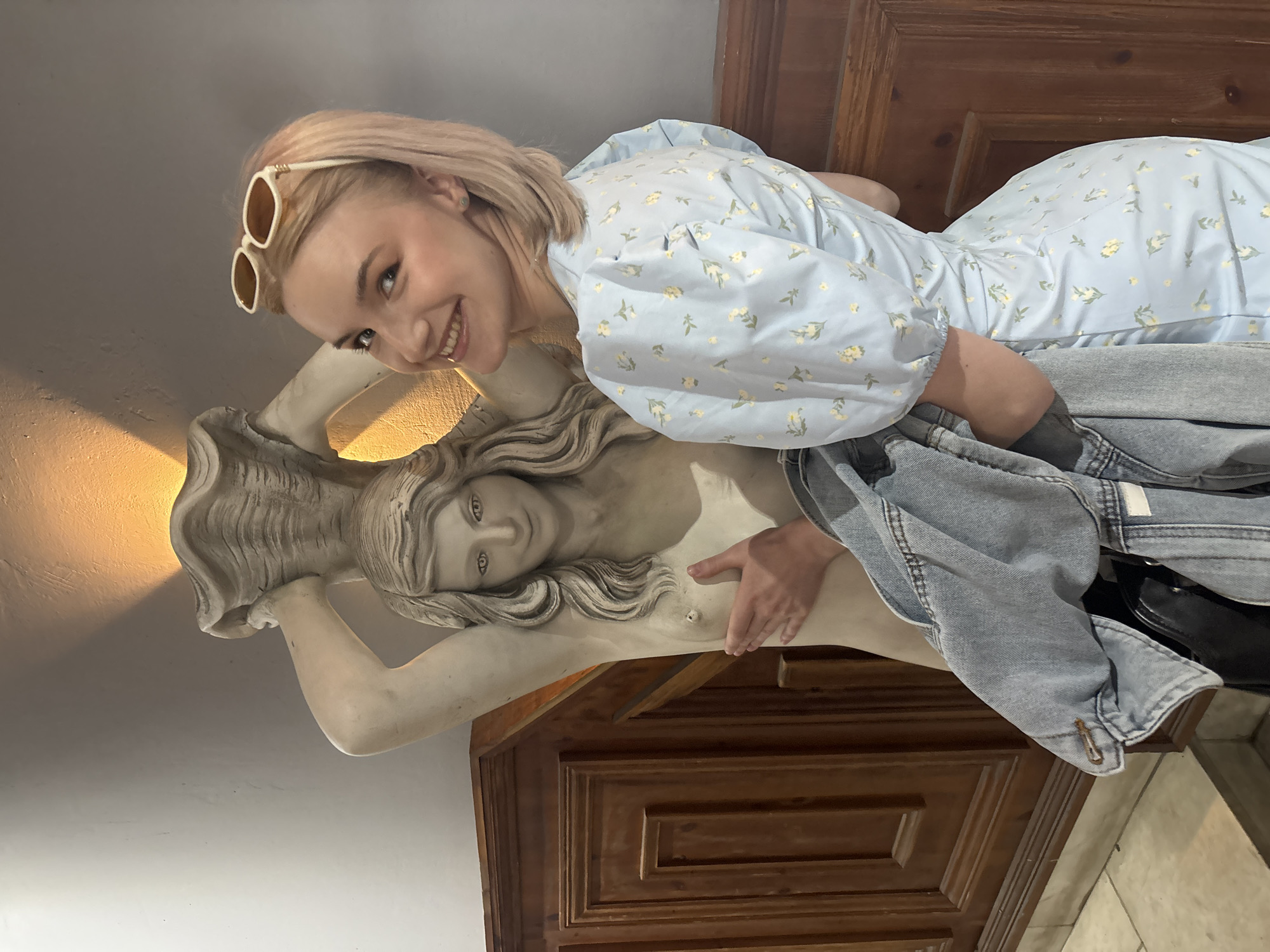 An indoor, eye-level photograph taken in Saint Petersburg, Russia, depicts a young blonde Caucasian woman posing playfully next to a classical-style, light-colored statue of a nude female figure. The woman, smiling broadly at the camera, wears a light blue dress adorned with small yellow floral patterns and puffy sleeves. Cream-colored sunglasses are pushed up onto her head. Her right hand is positioned to cover the left breast of the statue, while her left arm holds a light blue denim jacket. The statue, possibly made of marble or plaster, features a female figure with long, flowing hair holding a large conch shell above its head, which appears to house a light source casting a warm orange glow onto the light gray wall above it. The background consists of dark brown, intricately carved wooden paneling.