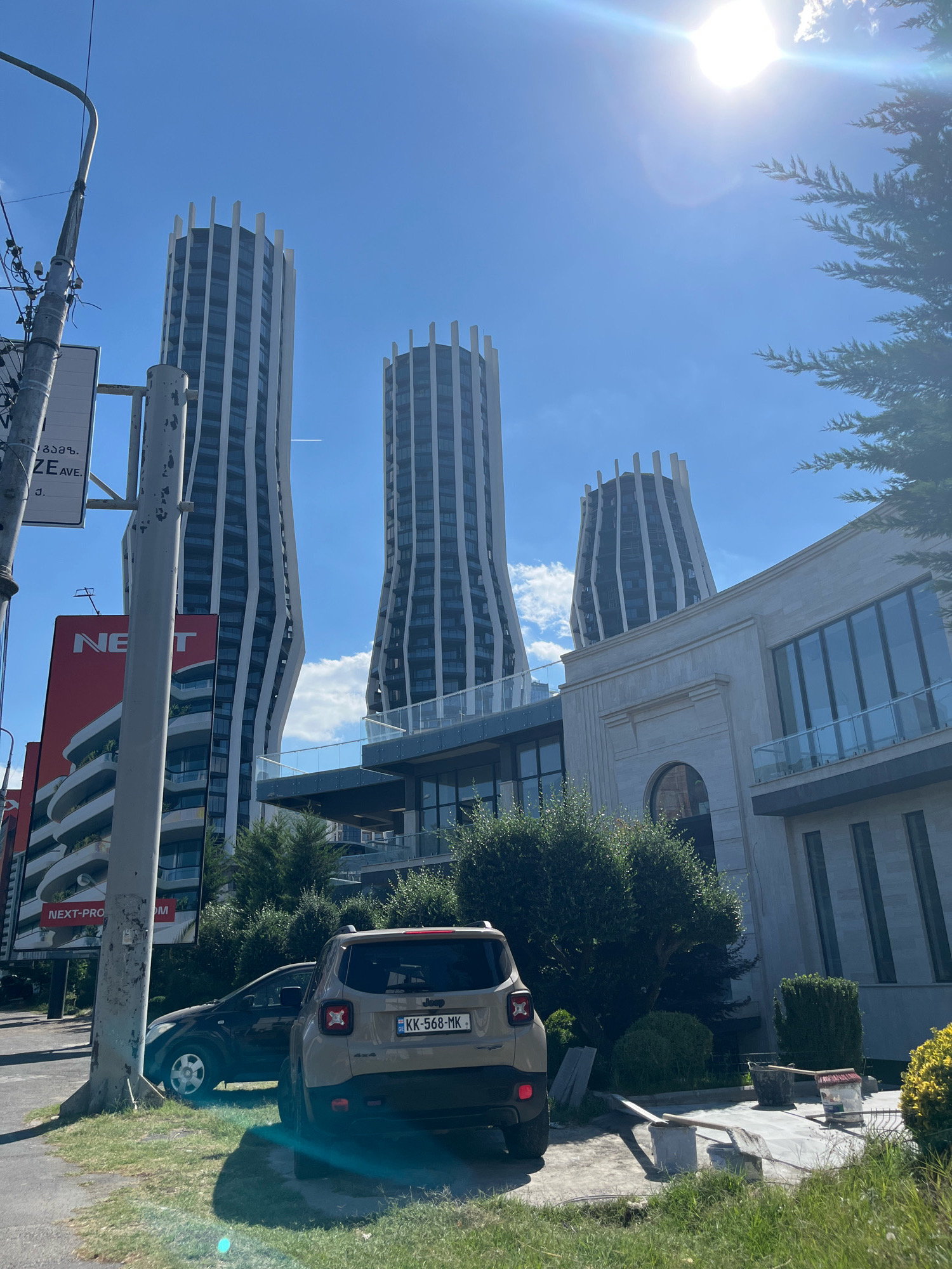In Tbilisi, Georgia, under a bright sun and clear blue sky, a street scene unfolds. Three striking, modern high-rise buildings with unique, curved facades dominate the background. Their architecture features vertical white fins against dark windows. In the foreground, a beige Jeep Renegade is parked on the right side of the frame. A dark sedan is parked behind it. A grey pole with electrical wires and street signs stands prominently on the left. A modern, light-colored building with large windows and stone accents is visible in the mid-ground. Greenery, including trees and bushes, surrounds the buildings and cars, suggesting a well-maintained urban landscape. Construction materials, such as buckets and shovels, are scattered on a paved area to the right, indicating ongoing work. The overall impression is a blend of contemporary architecture and everyday urban life in Tbilisi.