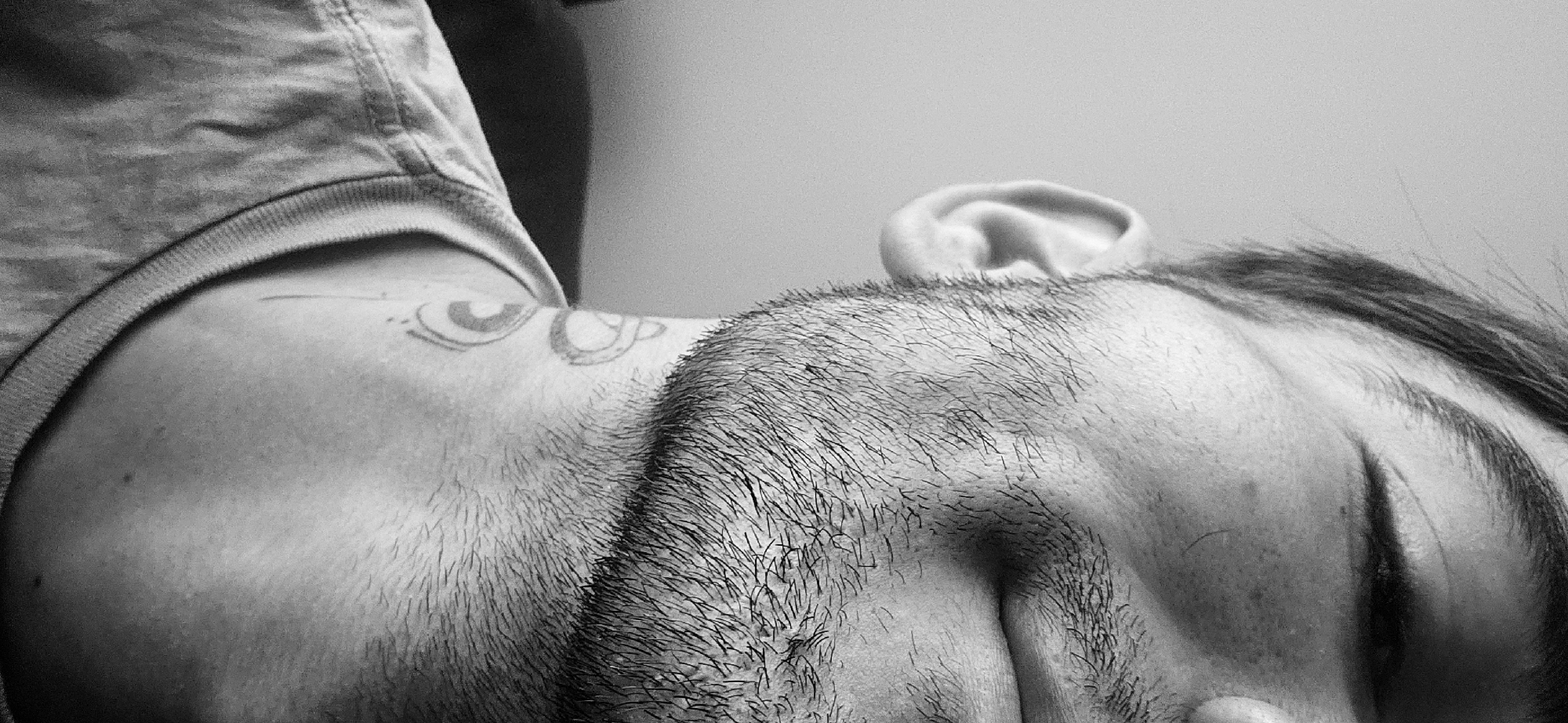 A black and white close-up photograph shows a man lying on his side, facing away from the viewer. His head rests on what appears to be his hand. The man has short, dark hair and a beard. Part of his ear is visible near the top of the frame. He is wearing a t-shirt and there is a tattoo on his shoulder. The background is a plain wall. The image provides no information regarding the location in Plovdiv, Bulgaria.