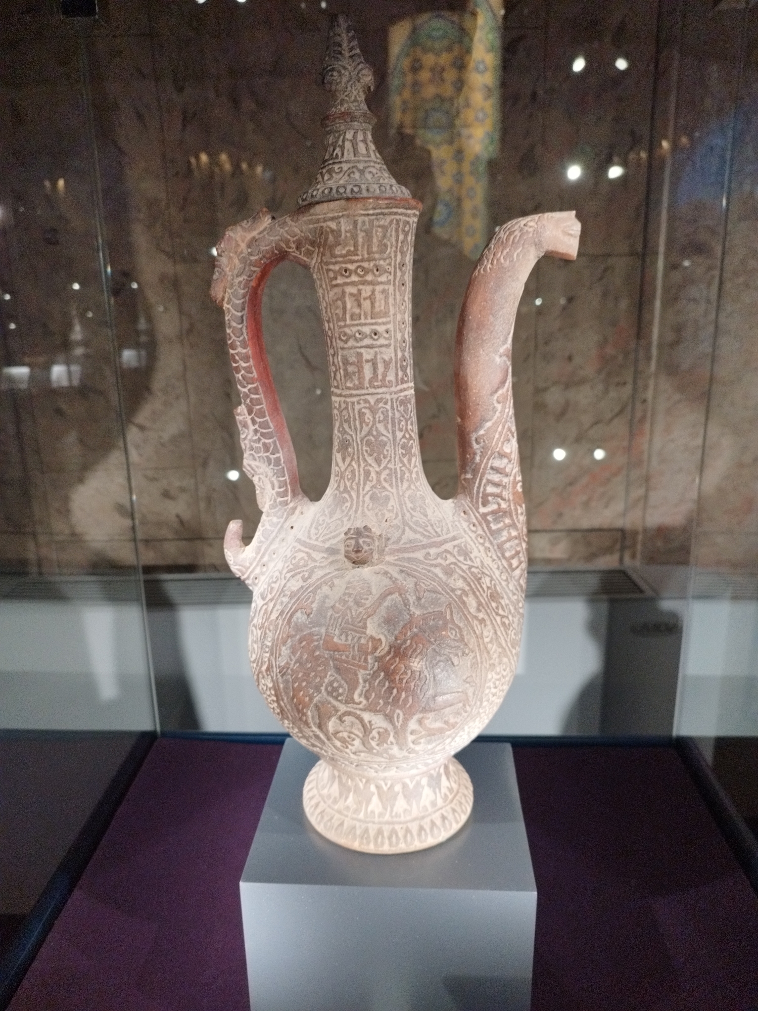 A close-up, eye-level, full shot shows an ornate, terracotta jug with a thin, long spout, and a curved handle. The jug is decorated with intricate patterns and imagery, including what appears to be a human figure in a circular motif on the body, and detailed relief carvings. The handle is shaped like a dragon or serpent, with scales and a head at the end. The jug has a flared base with a band of repeating figures. It is displayed on a light gray pedestal, which is on top of a dark purple surface. The jug is enclosed in a glass display case, with reflections of lights and the environment visible on the glass. The background is blurred, suggesting a museum or exhibition setting. The jug's texture is rough and appears to be ancient.