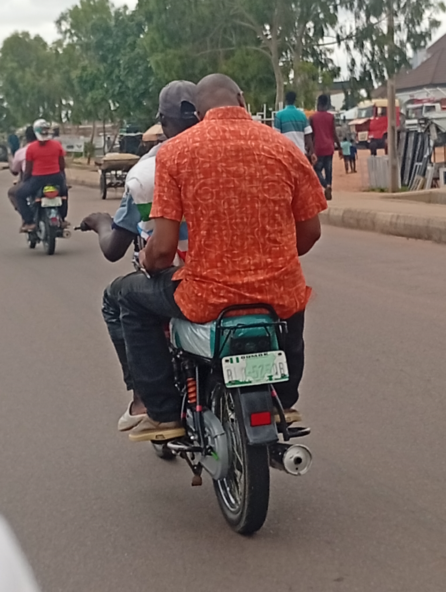 In Tumfure, Nigeria, two individuals are riding on a motorcycle down a paved road. The rider in the front, wearing a grey baseball cap and a blue and white striped shirt, has his left hand on the handlebars. The passenger in the back is wearing an orange, patterned short-sleeved shirt and dark pants. The motorcycle is black with a green seat and has a license plate that reads "RU-575-DR". In the background, trees line the side of the road, and various buildings and other vehicles, including a cart and other motorcycles, are visible. The sky is overcast.