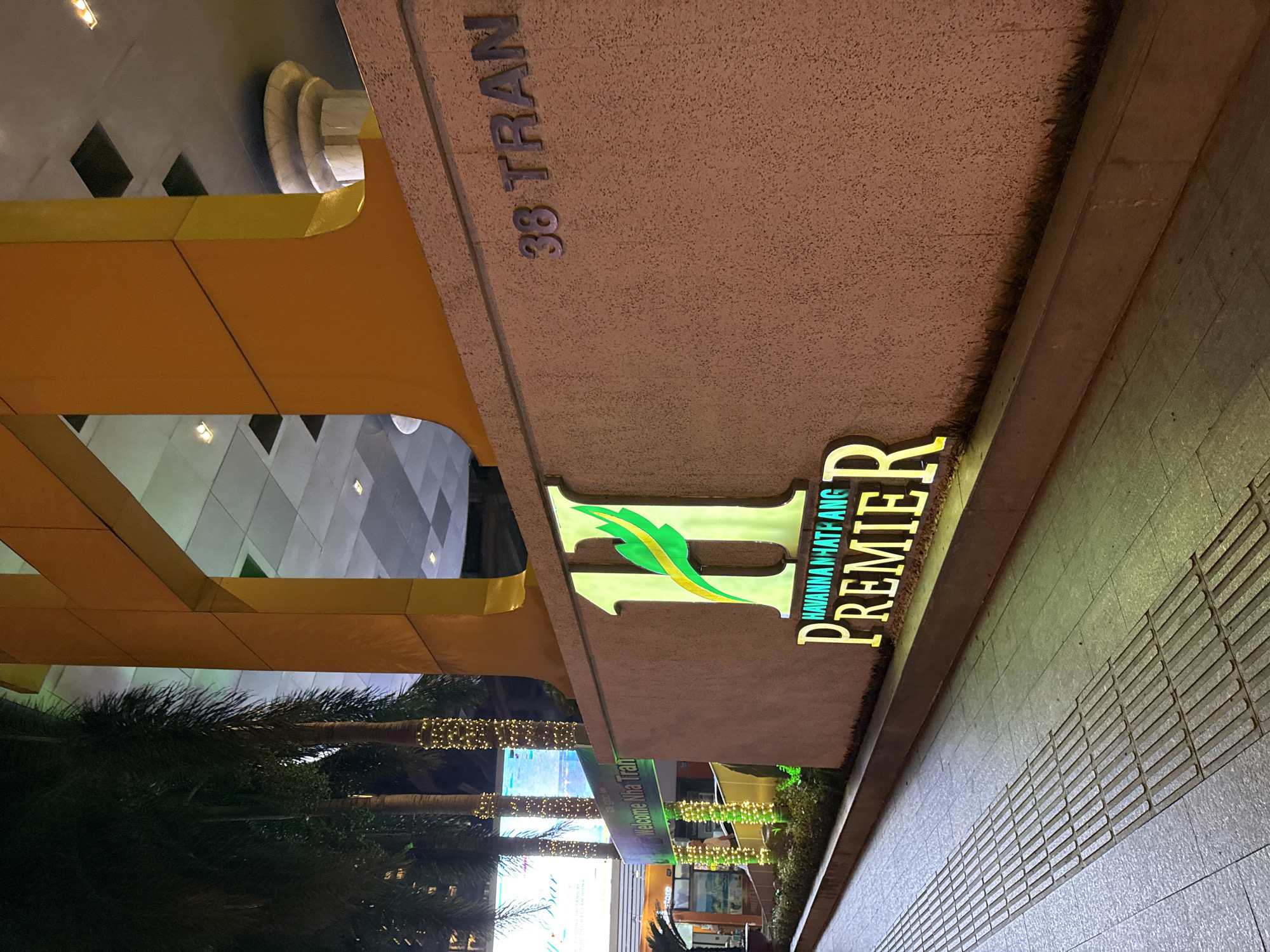 The image shows the facade of a building at night in Nha Trang, Vietnam. A large, illuminated "H" logo with a green leaf design is prominently displayed on a textured, tan-colored wall. Below the "H" logo, the words "HAVANNA NHA TRANG PREMIER" are also illuminated. To the right of the logo, the address "38 TRAN" is visible, also in illuminated lettering. Palm trees decorated with string lights are situated to the left of the building facade. In the background, a large screen displays text, and part of a building with yellow and grey elements is visible. A tactile paving path runs along the sidewalk in the foreground. The overall scene suggests a commercial or hospitality establishment.