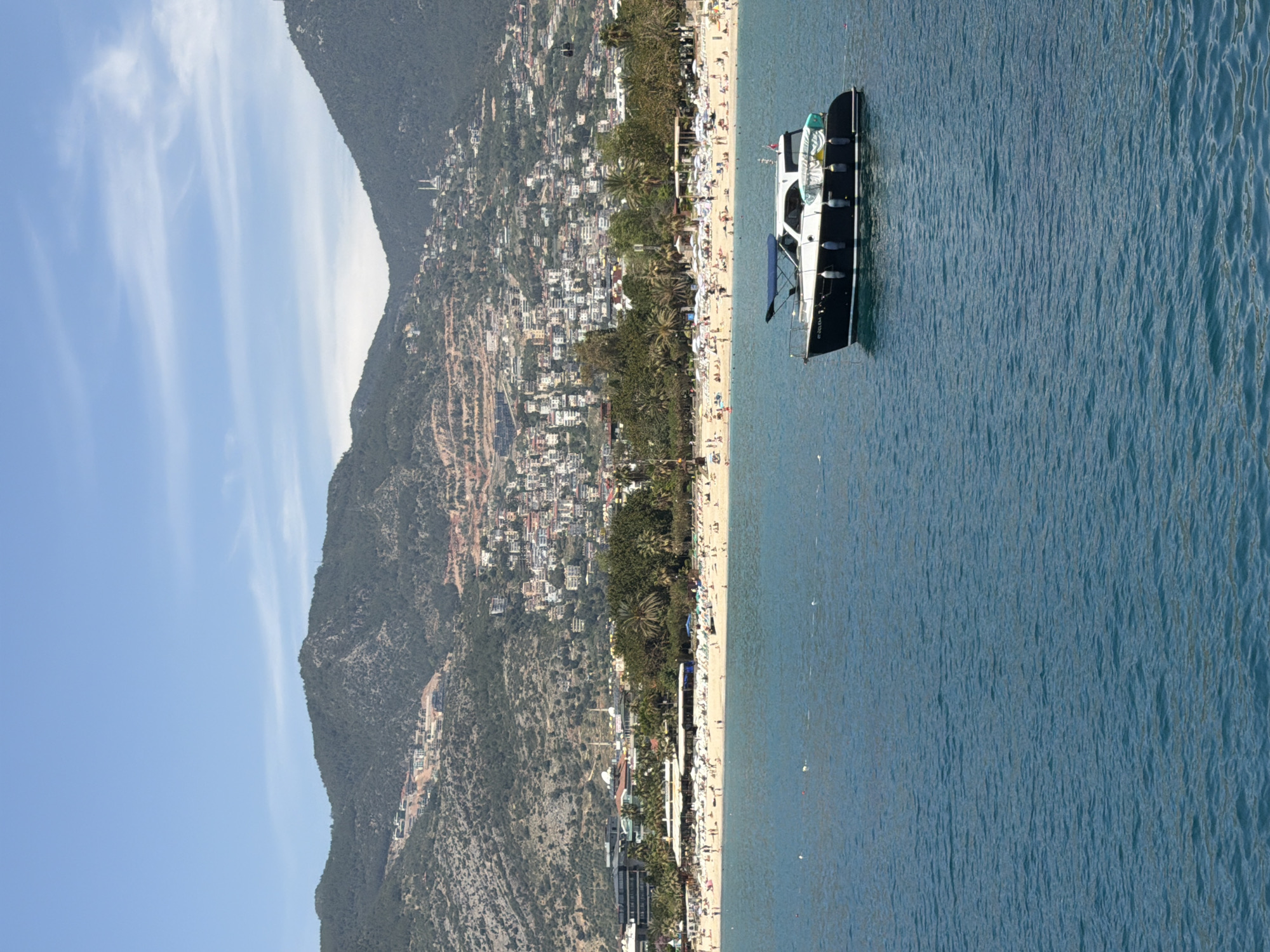 The image captures a coastal scene in Alanya, Turkey, featuring a beach, a mountain range, and a body of water. The composition is oriented vertically.
On the right side, a blue body of water occupies a significant portion of the image. A black and white boat is docked in the water near the coastline.
Adjacent to the water is a sandy beach densely populated with people. Beach umbrellas and sunbathers are visible.
Along the coastline, various buildings and structures are present. Beyond the immediate coastline, a mountain range rises, and the slopes of the mountain are dotted with vegetation and buildings.
The sky is partially visible on the left side of the image, showcasing a blue expanse with some wispy clouds.