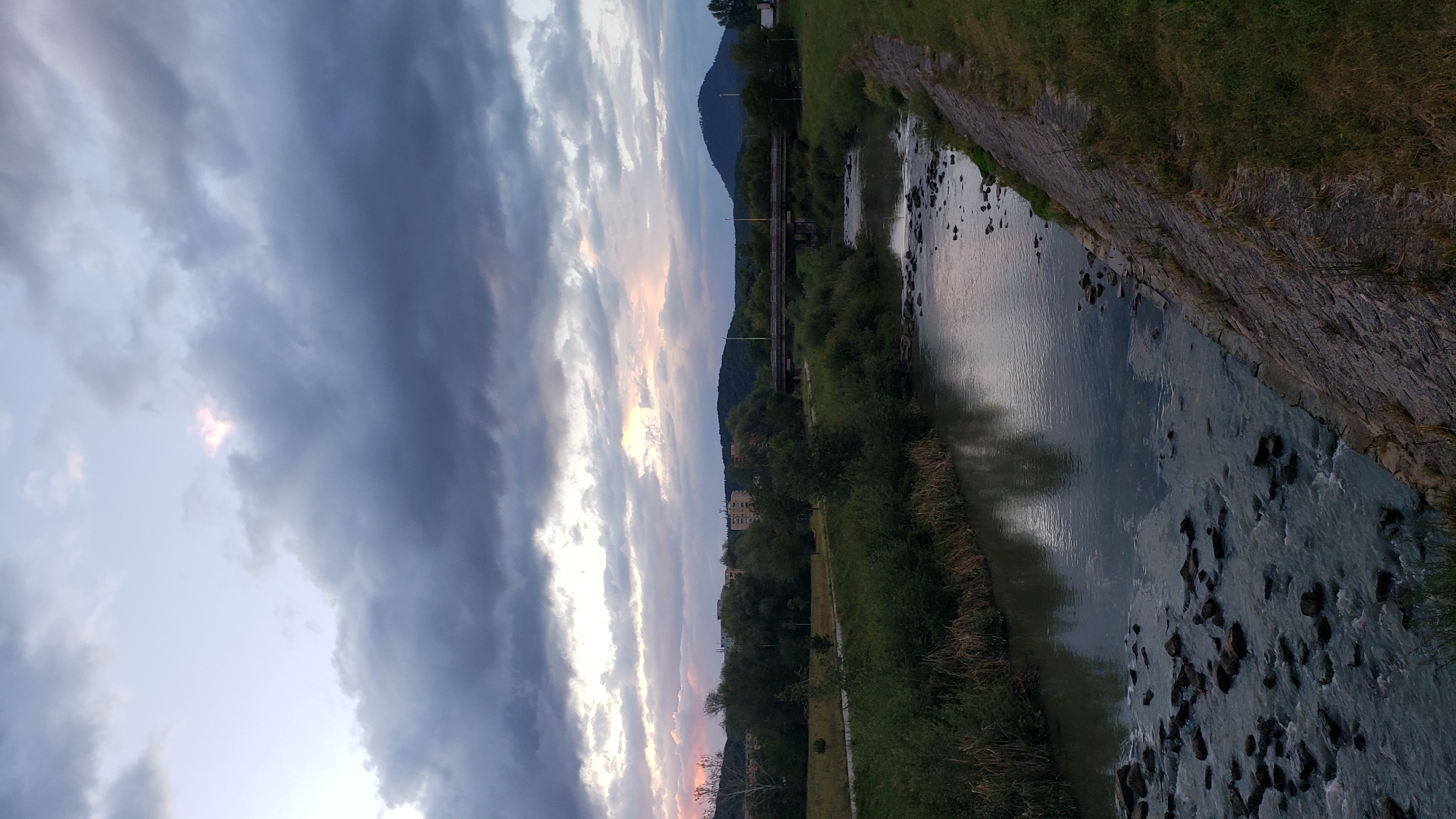 The image depicts a river flowing through an urban landscape at dusk. The sky is filled with dramatic, dark clouds, with streaks of pink and orange visible near the horizon, suggesting the sun has recently set. A bridge spans the river in the middle ground. On the left bank of the river, there are trees and buildings, indicative of a city. The right bank is covered in grass and a stone embankment. The river itself is shallow, with rocks visible beneath the surface and a reflection of the sky on its water. This scene is located in the city of Prešov, Slovakia.