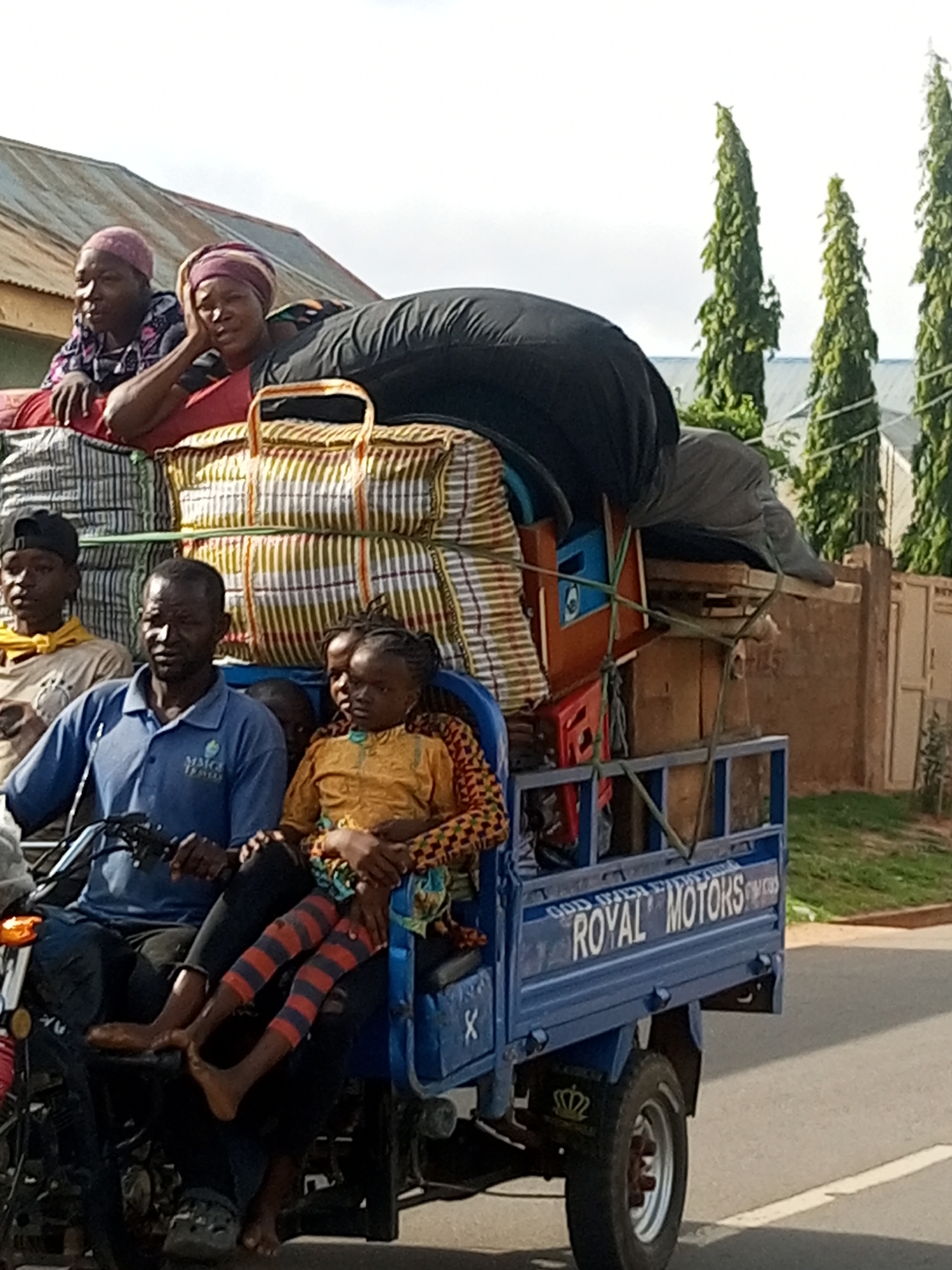 A three-wheel cargo motorcycle transports several people and their belongings through Tempure, Nigeria. Two women, wearing headscarves, sit at the front of the motorcycle on a stack of luggage. Behind them, a man drives the motorcycle. In front of him, a young girl in a yellow shirt and a child in a patterned outfit are seated. Several large bags and boxes are piled on the back of the motorcycle, secured with ropes. The motorcycle is branded with "ROYAL MOTORS" in white lettering. The background features buildings and tall, slender trees. The sky is overcast.