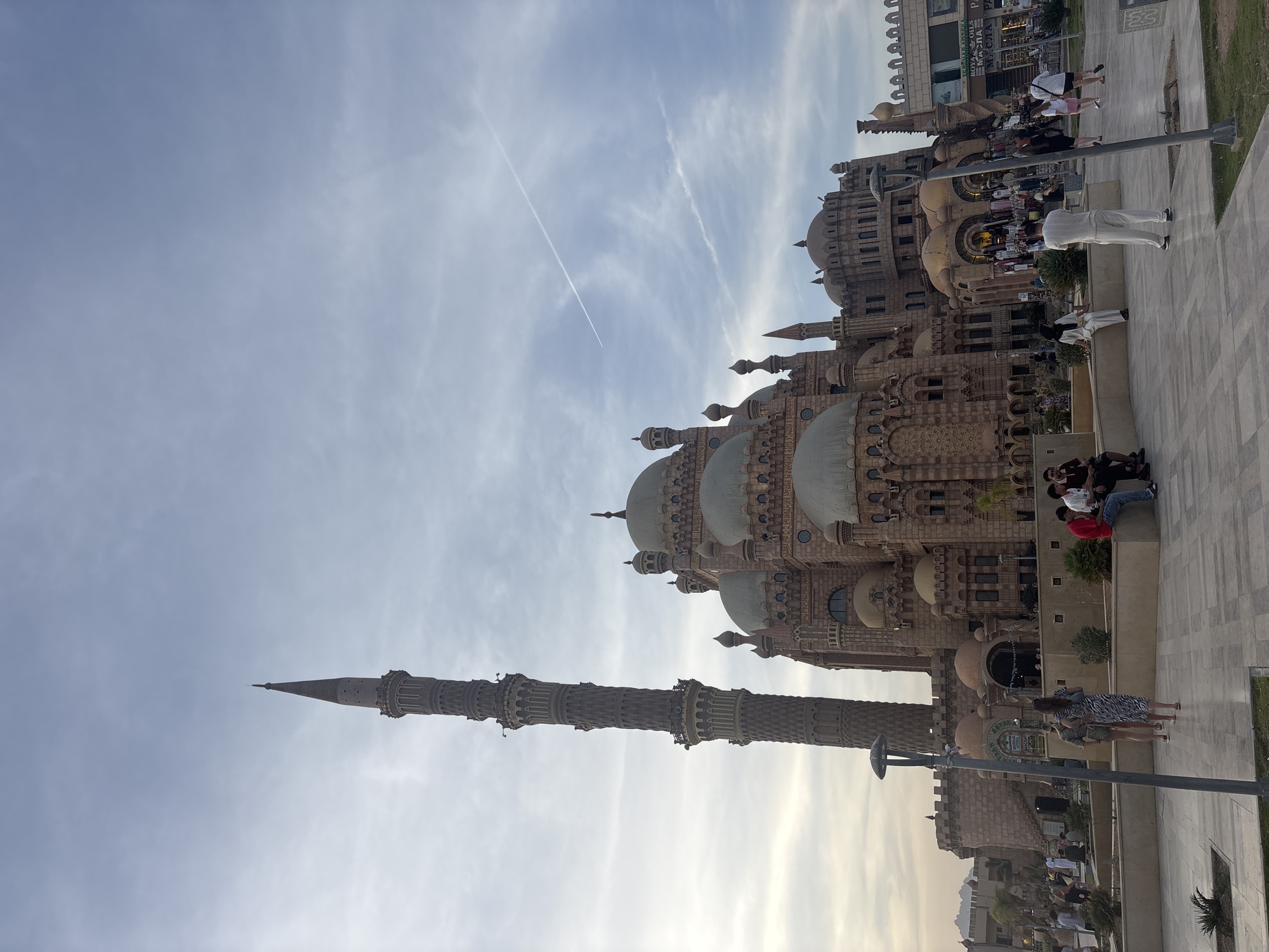 The image depicts a large mosque complex under a partly cloudy sky with a contrail. The mosque is constructed of light brown stone and features multiple domes and a tall minaret. People are present in front of the mosque, including individuals sitting on a low wall and others walking nearby. Commercial buildings are visible in the background. The city location is not identifiable from the image.