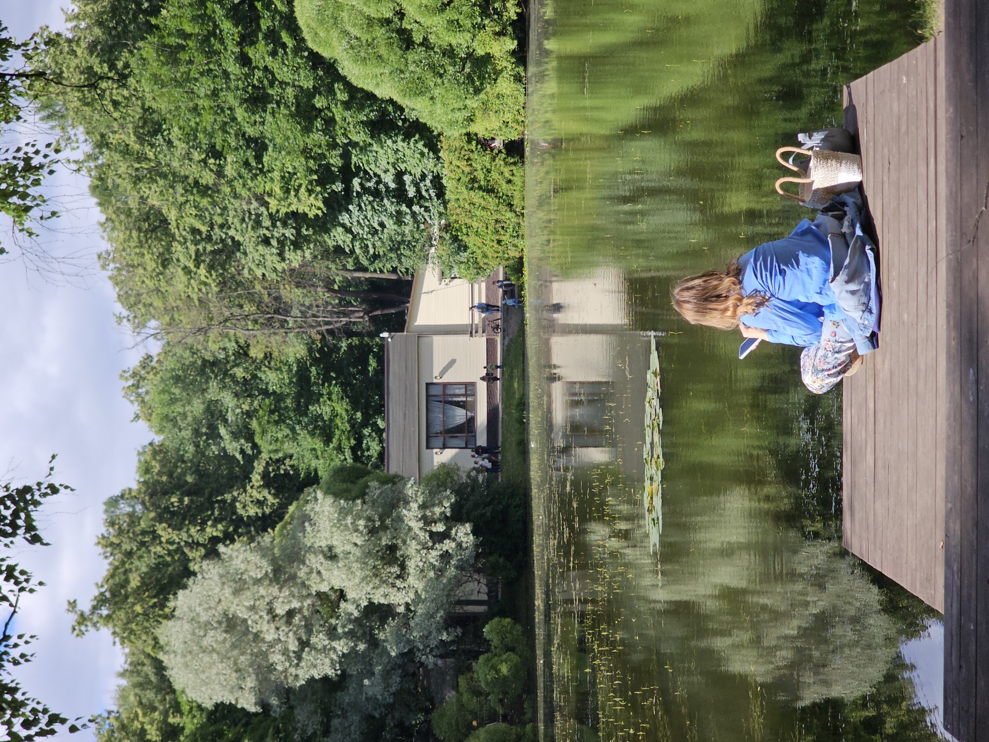 A woman is sitting on a wooden pier facing a lake in a park. She is wearing a blue shirt and patterned pants and is looking at her phone. Next to her are two woven bags. The lake is calm and reflects the surrounding trees. In the background, there is a building with windows and several people walking. The scene appears to be a peaceful day in a city park, possibly in Moscow, Russia, given the architectural style and vegetation.