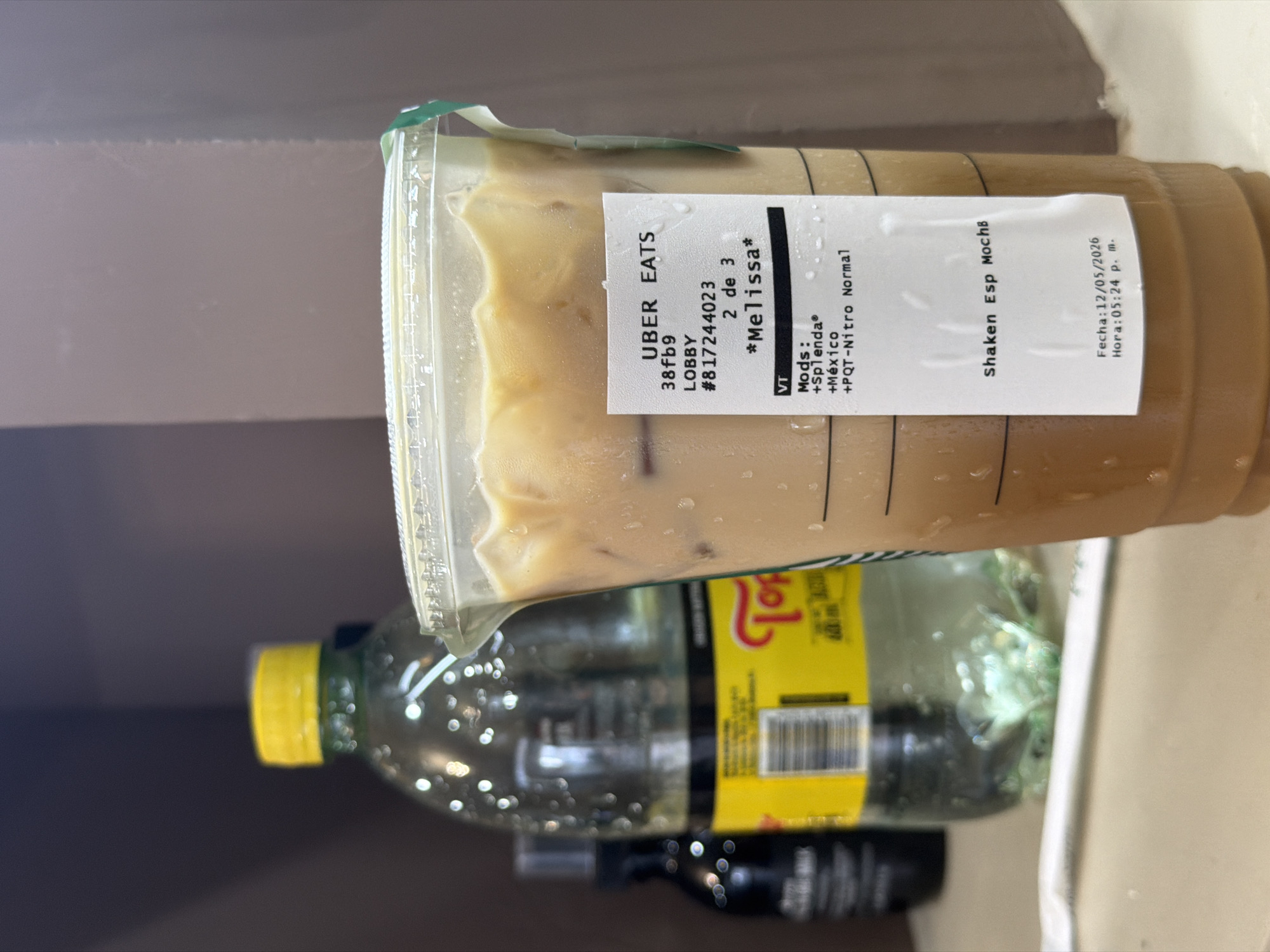 A Starbucks Shaken Espresso Mocha drink from Uber Eats is next to a bottle of Topo Chico sparkling water. The Uber Eats order was picked up by the driver in the lobby on May 12, 2026 at 5:24 p.m. for a customer named Melissa. The order was two out of three items purchased. The drink contains Splenda, a sweetener, and PQT-Nitro Normal. There is also a black bottle in the background. The setting looks like a kitchen counter or sink. The drinks were presumably purchased in Villahermosa, Mexico.