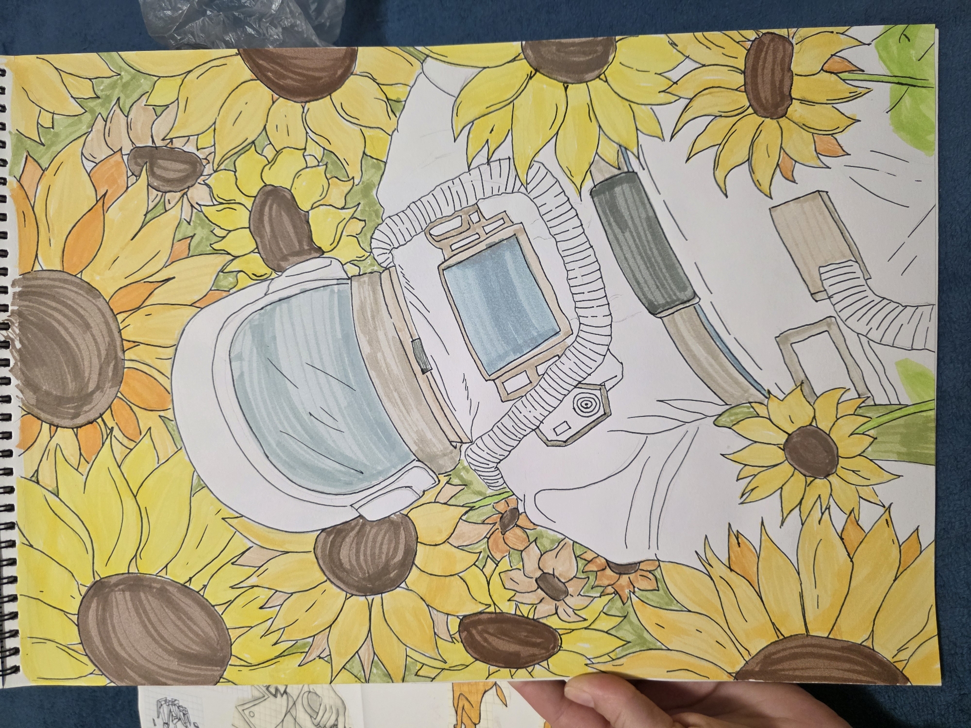 A colored drawing on paper depicts an astronaut in a spacesuit lying among numerous sunflowers. The astronaut's helmet, predominantly white with a blue visor, is positioned towards the upper left, while the rest of the body extends across the page. The suit includes details like ribbed tubing and control panels rendered in shades of grey and brown. The sunflowers have yellow petals and brown centers, creating a vibrant background. The drawing is on a spiral-bound notebook, and a hand holds the bottom right corner of the page. A blue surface is visible in the background, and part of a plastic bag is at the top. Below the drawing, a section of another drawing is visible.