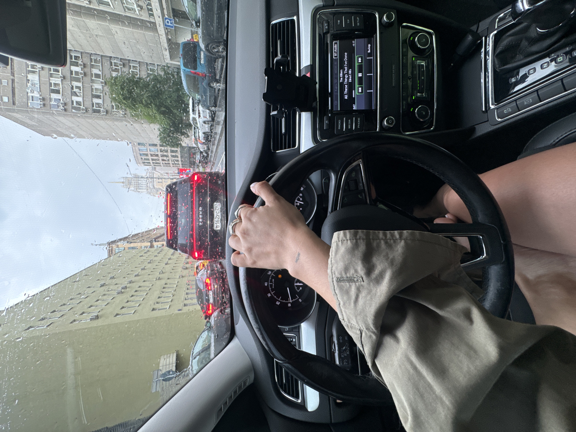 The image depicts a first-person perspective from the driver's seat of a car stopped in moderate traffic on a rainy day in Moscow, Russia. The car's interior is visible, with a hand on the steering wheel. The dashboard displays a car stereo system playing music by "The Killers." The windshield is wet with raindrops, and the cityscape outside shows apartment buildings and other vehicles. The road appears wet and reflective, characteristic of rainy conditions in an urban environment. The sky is overcast.