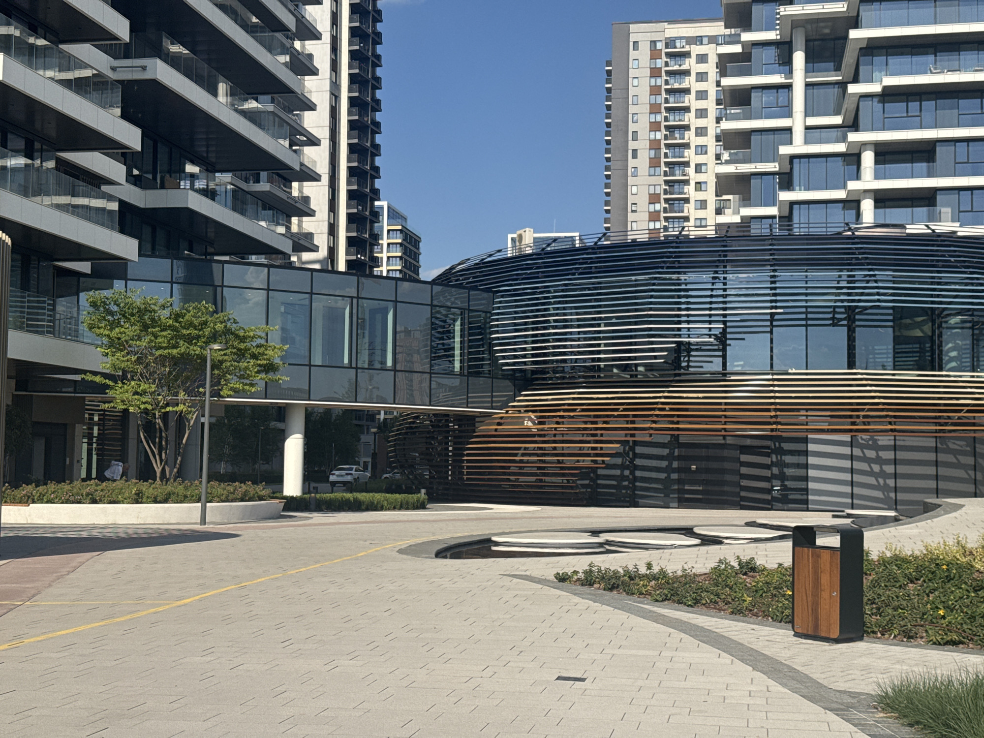 This media file depicts an exterior view of a modern urban complex on a bright, sunny day. The scene is dominated by contemporary architecture, featuring tall residential buildings with repeating balconies and a unique, curved structure with horizontal slatted elements and reflective glass. A glass-enclosed walkway connects two of the buildings.
In the foreground, a paved courtyard with patterned tiles and yellow lines serves as a pathway. A dark trash receptacle with a wooden front is situated near a landscaped area with low green bushes and some yellow flowers. A light-colored car is visible parked in the middle ground. A young tree with lush green foliage stands near one of the buildings, casting a shadow on the pavement. A person in white clothing can be seen through the glass of a building entrance.
The sky is a clear, vibrant blue, suggesting fair weather and midday. The location cues, including architectural style and the specific mention of Belgrade, Serbia, place the scene within a modern development in that city. There is no visible text in the image. The overall impression is one of a clean, well-maintained, and contemporary urban environment.