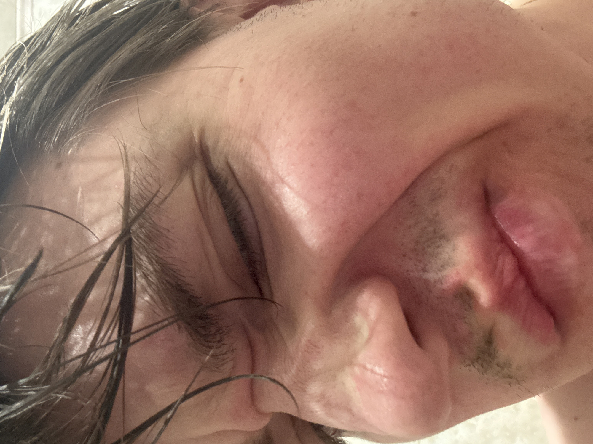 This is a close-up, eye-level, indoor, medium shot of a person's face, partially obscured by dark, wet hair. The individual is looking down and to the left, with their eyes closed or squinting. The skin is pink and appears to be flushed, particularly on the cheeks. There are visible wrinkles around the eye and on the cheek. The lips are slightly parted and appear red. The person has some stubble on their upper lip and chin. The background is a blurred, light-colored surface, possibly a wall or a textured fabric. The image does not provide any information about the location in the city of Dnipro, Ukraine.