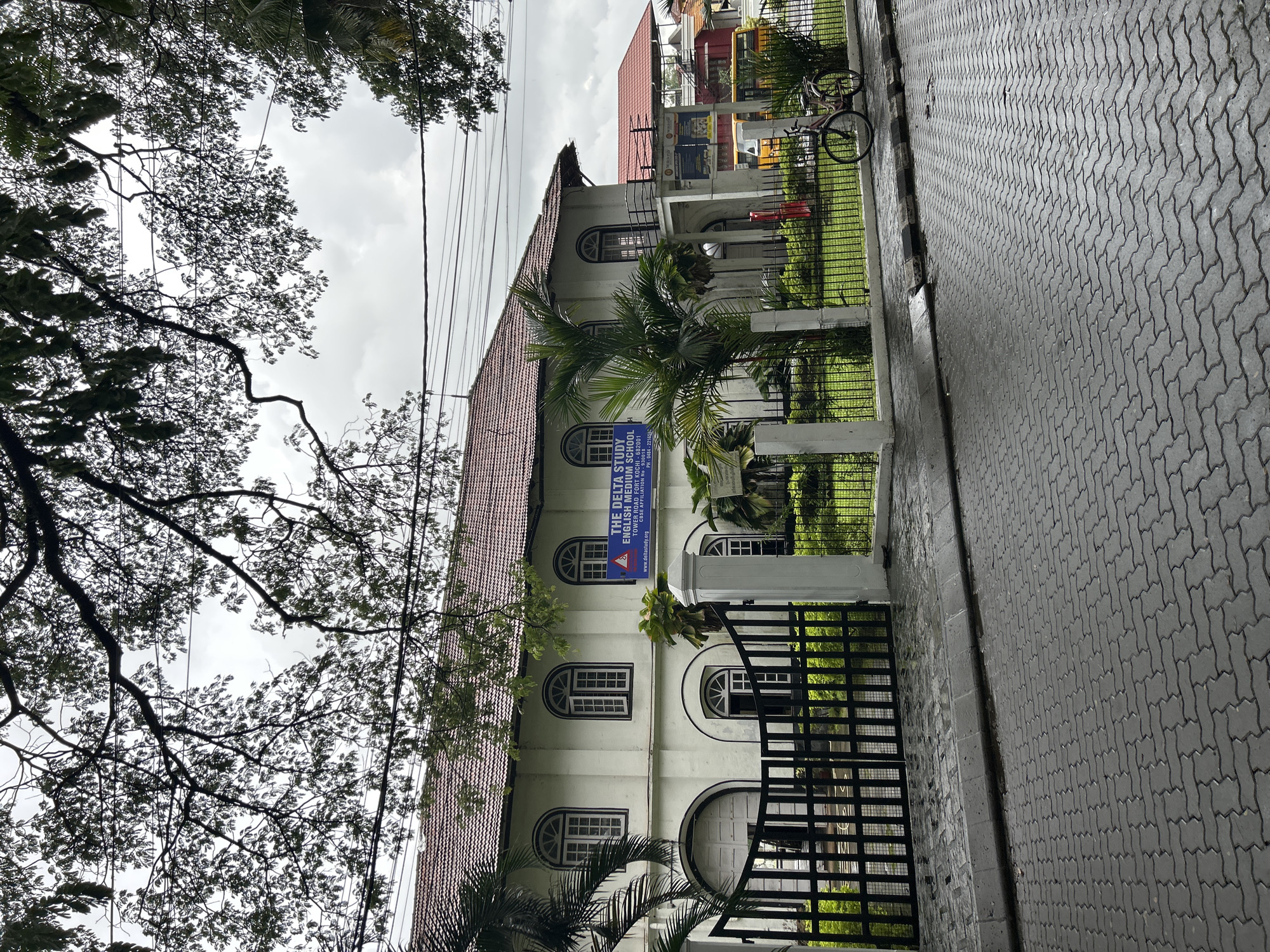 The image displays an exterior view of "THE DELTA STUDY ENGLISH MEDIUM SCHOOL" in Kochi, India, captured on an overcast day. The central subject is a multi-story, light-colored building featuring a red-tiled roof and multiple arched windows with white frames. A prominent blue rectangular sign, partially obscured by a palm tree, is affixed to the building. The sign clearly states "THE DELTA STUDY ENGLISH MEDIUM SCHOOL," along with its address: "TOWER ROAD FORT KOCHI - 682001," a "CBSE AFFILIATION No - 930018," a phone number "PH-0484-221456," and its website "www.deltastudy.org."
In front of the school, a black metal fence and gate enclose a green lawn. A bicycle is parked behind this fence on the right side of the frame. Adjacent to the fence, a grey paved pathway made of interlocking blocks runs along the foreground, appearing somewhat damp, suggesting recent rain or high humidity. The sky is largely overcast with light grey clouds. Overhead, numerous black power lines crisscross the upper portion of the scene. Lush green trees with dense foliage and some prominent branches frame the left and top areas of the image, contributing to the tropical setting typical of Kochi. No people or specific activities are visible, presenting a quiet street scene during daylight hours.