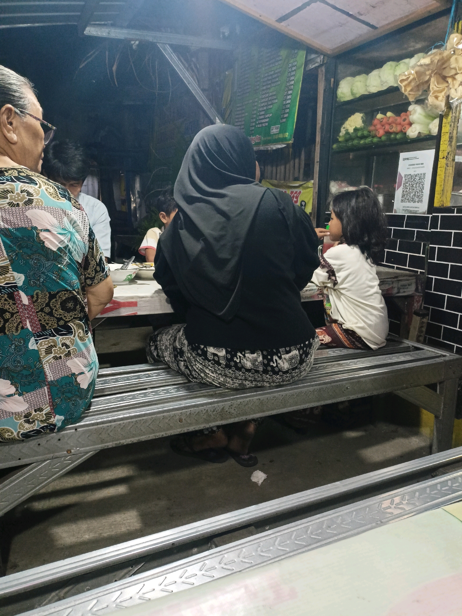 An overhead view shows a group of people eating at a food stall in Surabaya, Indonesia. A woman wearing a black hijab sits at a table, her back to the camera, while a young girl in a white shirt and patterned leggings eats beside her. To the left, an older woman with glasses and a colorful patterned shirt also sits at the table, facing away from the camera. Behind them, a man in a white shirt is visible. The food stall displays various vegetables and packaged snacks, including lettuce, tomatoes, cauliflower, and crackers, on shelves. A QR code is displayed on the wall. The overall scene depicts a casual dining experience at a local eatery.