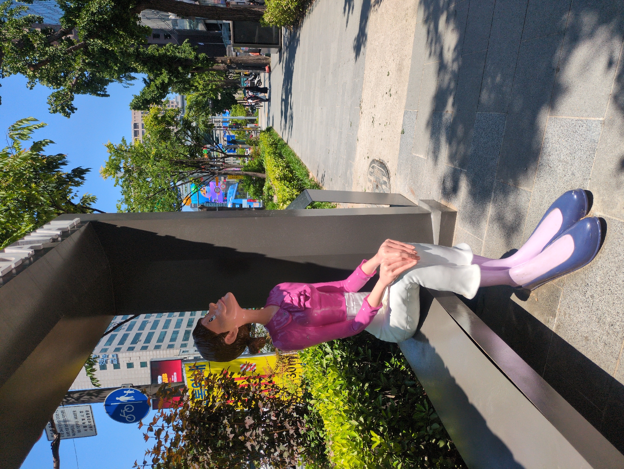 The image shows a seated, stylized statue of a woman. She is wearing a pink top, white pants, and purple and light purple shoes. The statue is seated on a low concrete structure, possibly part of a bus stop or shelter. Vegetation is located behind the statue.
In the background, a city street scene is visible with trees, buildings, and street signs. A blue sign showing the symbol of people and bicycle indicates a pedestrian or bicycle path. Based on the Korean text on the signs and the appearance of the streetscape, this image is likely taken in Seoul, South Korea.