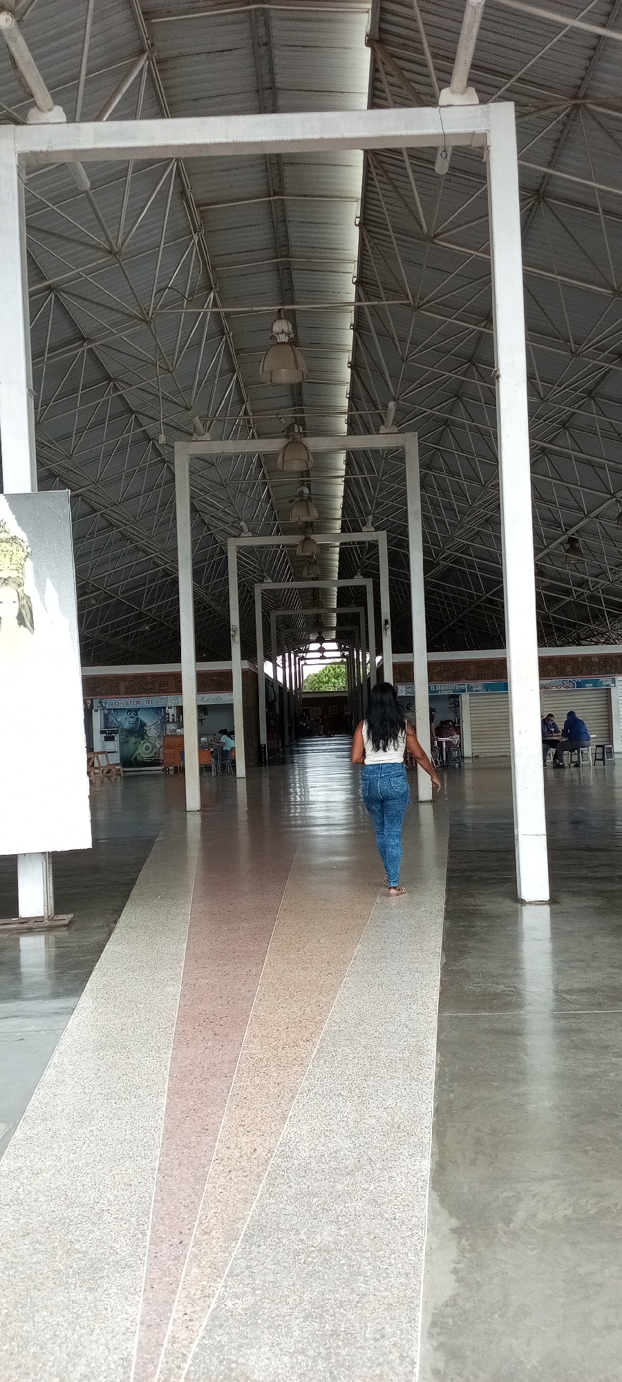 The image depicts a woman walking away from the viewer in a large, covered market or hall. The woman is wearing a white top and blue jeans. Overhead, there is a metal roof structure supported by white pillars and beams that form a series of archways. The floor has a patterned walkway with alternating light-colored sections. There are stalls or shops along the sides of the walkway with some people sitting at tables. The location appears to be an open-air marketplace.