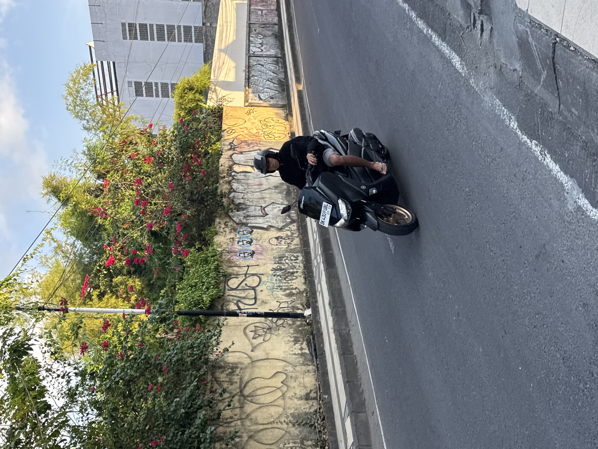 This daytime image captures a street scene in what appears to be an urban or semi-urban area, consistent with Kuta, Indonesia. A person is seated on a black scooter, which is parked or stopped at the side of the road, next to a concrete curb. The individual wears a black long-sleeved top, shorts, and a light-colored helmet, with their feet bare. The scooter features a gold-colored wheel and its license plate prominently displays "DK 4309 UBL" and "07 28".
Adjacent to the scooter, a low wall runs parallel to the street, heavily adorned with various graffiti tags, shapes, and what look like abstract faces. Partially legible text, such as "HUSTLE", can be made out. This wall is partially obscured by lush green bushes and vines that are vibrant with clusters of red or pink flowers, likely bougainvillea. Further back, more dense green foliage and trees are visible, with power lines passing through. In the background, a light-colored, multi-story building with numerous small, dark windows stands under a clear blue sky with scattered white clouds.
The road surface is asphalt, with a distinct white line marking the edge of the travel lane near the curb. The lighting suggests it is daytime, possibly morning or late afternoon, characterized by bright light and soft shadows. The overall scene conveys a casual, everyday moment in a tropical setting.