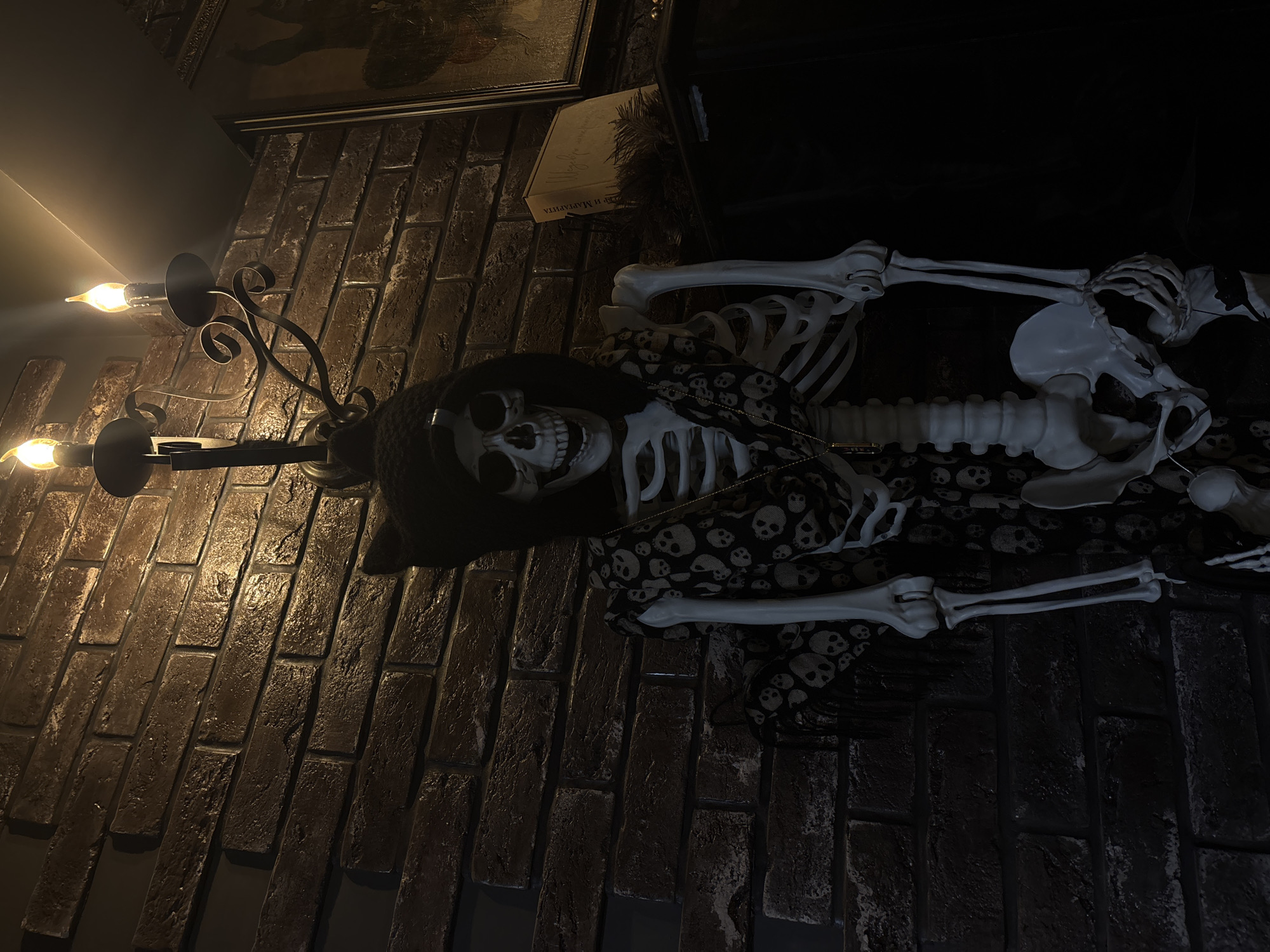 The image shows a Halloween-themed display on a brick wall. A skeleton, dressed in a hooded cloak and a fabric printed with skulls, is draped over the wall. A candelabra with two lit bulbs is mounted on the wall to the left of the skeleton. A framed picture is visible on the wall above the skeleton and candelabra. The scene is dimly lit, with the light from the candelabra illuminating the brickwork and parts of the skeleton. The overall mood is eerie and festive, consistent with Halloween decorations. The location is identified as Sosnovo, Russia. No visible text is present in the image.