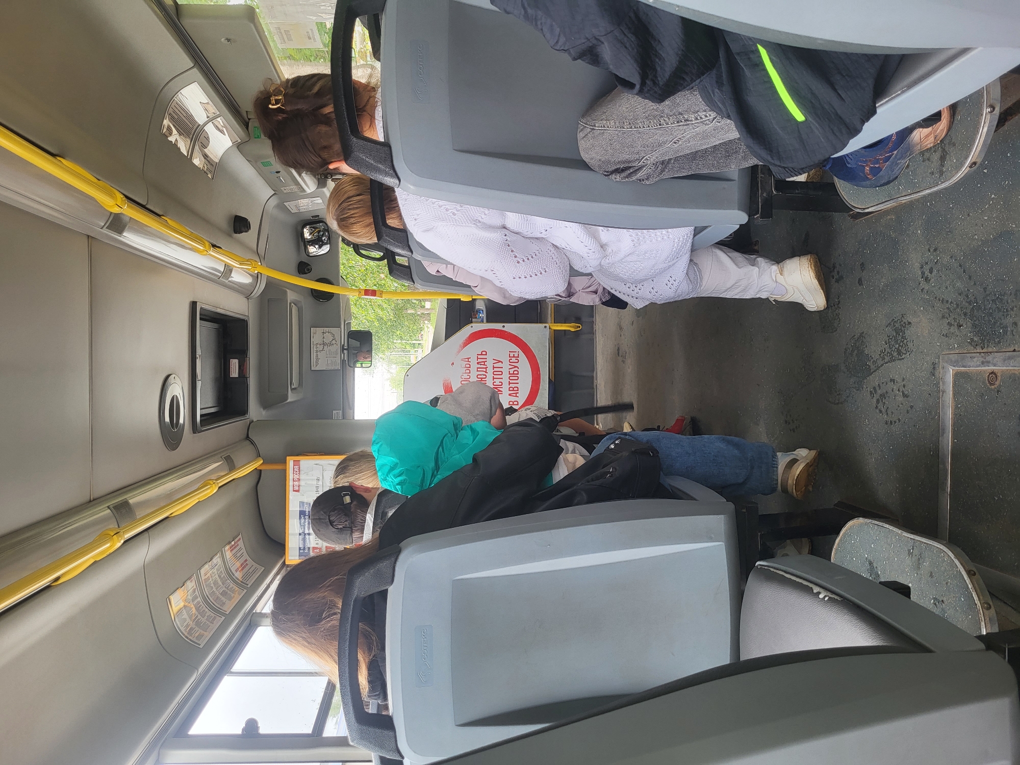 The image captures the interior of a public transit bus in Chaikovsky, Russia, oriented sideways, during bright daytime hours. Several passengers are seen from behind, occupying various seats within the vehicle.
The subjects include multiple individuals riding the bus, whose heads, shoulders, and backs are visible. Their attire is casual, including jackets, sweaters, jeans, and sneakers in various colors like black, white, turquoise, and grey. Objects visible are the bus seats, which are grey molded plastic with darker grey cushioned backs and seats. Yellow metal handrails extend along the ceiling and down vertical poles. Windows allow daylight to illuminate the interior, and the bus floor is a dark, somewhat worn material. A black bag is seen resting on one of the seats.
The activity taking place is simply passengers commuting or traveling on a bus. No specific events or interactions are clearly depicted; individuals appear to be quietly seated.
Notable details include the functional, somewhat industrial design of the bus interior. The bright lighting suggests it is daytime. Location cues are primarily given by the Cyrillic text visible on signs and posters. A prominent red and white circular sign clearly states: "ПРОСЬБА СОБЛЮДАТЬ ЧИСТОТУ В АВТОБУСЕ!", translating to "PLEASE OBSERVE CLEANLINESS IN THE BUS!". Other visible Cyrillic text fragments include "МОЯ РОССИЯ" ("MY RUSSIA"), "лтийский канал?" ("Baltic Channel?"), and "1848 году" ("in 1848"). The brand "SCOOTERC" is also visible on the back of a seat. Emotions of the passengers are not discernible from this perspective.