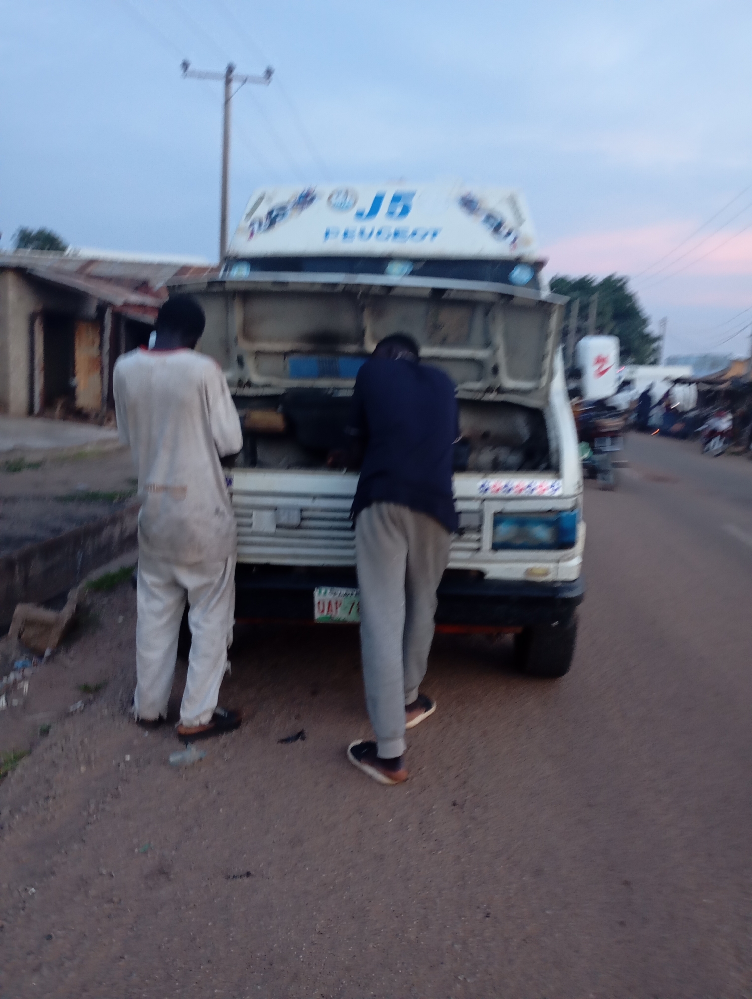 Two men are standing in front of a white van with its hood open on a dirt road in Shendam, Nigeria. The man on the left is wearing a white, long-sleeved shirt and matching pants. The man on the right is wearing a dark blue long-sleeved shirt and grey pants. The van has a "J5 PEUGEOT" logo on its side. A license plate with the characters "UAP 7" is visible on the front of the van. The background shows a building on the left and a street with other vehicles and people on the right. The sky is overcast with hints of pink and orange, suggesting it is either dawn or dusk. The overall impression is that the men are working on the van.